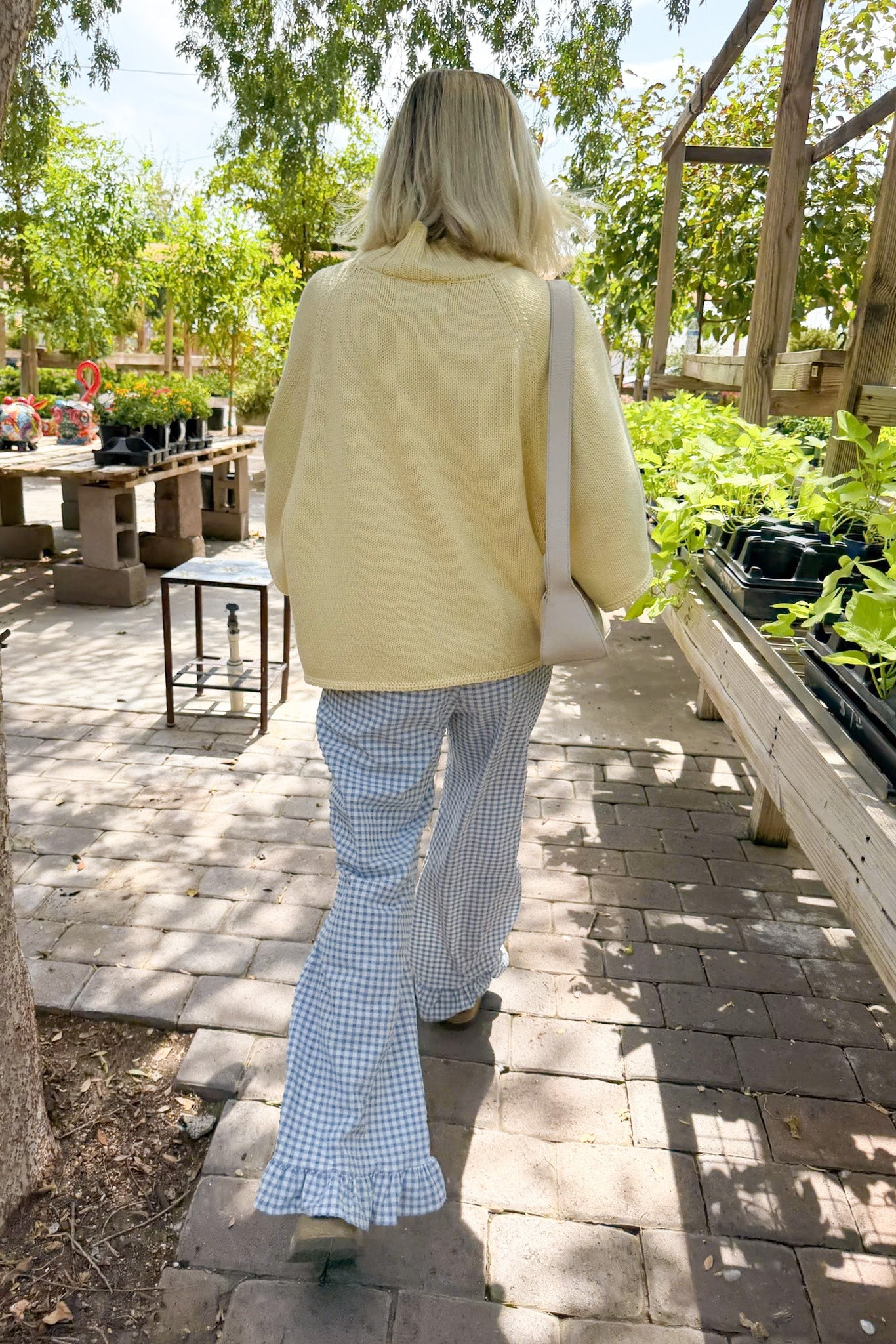 A woman viewed from behind walking on a paved path in a garden center, wearing a pale yellow knit sweater, blue-and-white gingham flared pants, and carrying a cream shoulder bag.