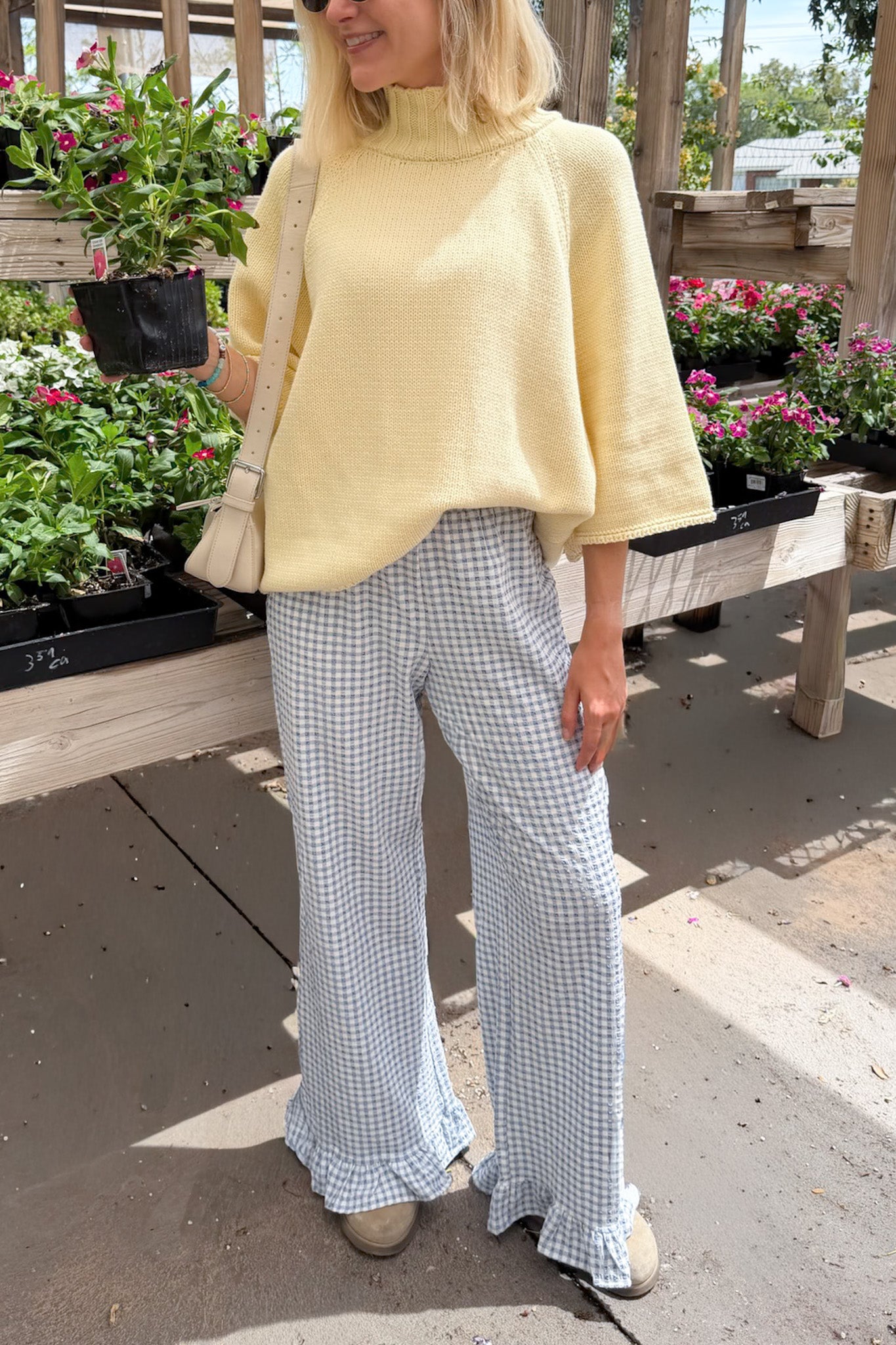 A woman wearing a pale yellow mock-neck knit top and light blue and white gingham wide-leg pants, standing in a garden center with potted plants in the background.
