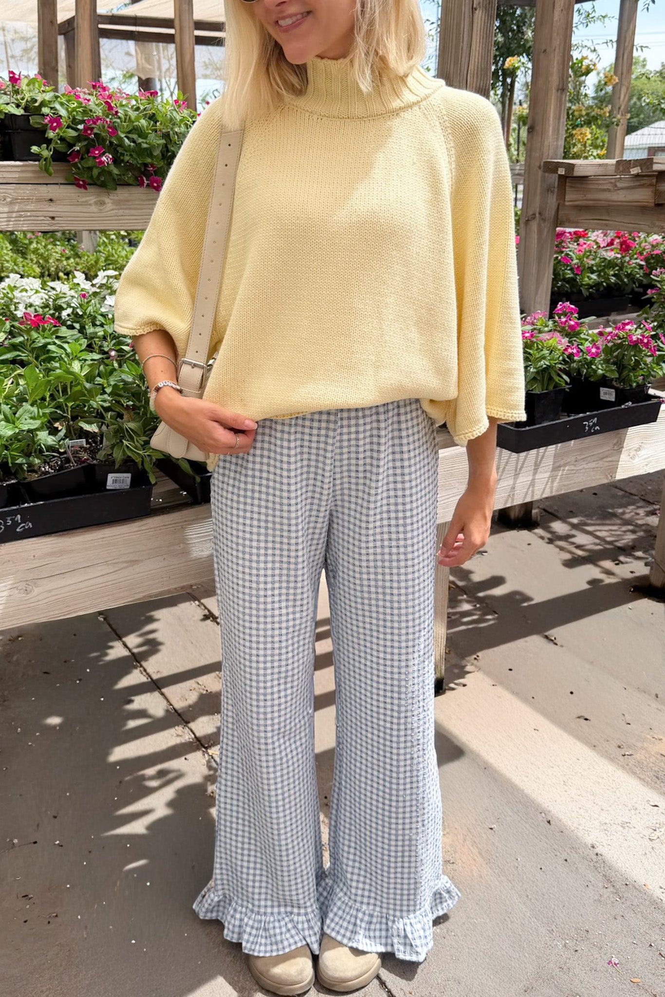 A woman wearing a pale yellow knit mock-neck tunic top and light blue and white gingham wide-leg pants with ruffled hems, standing in a garden center.