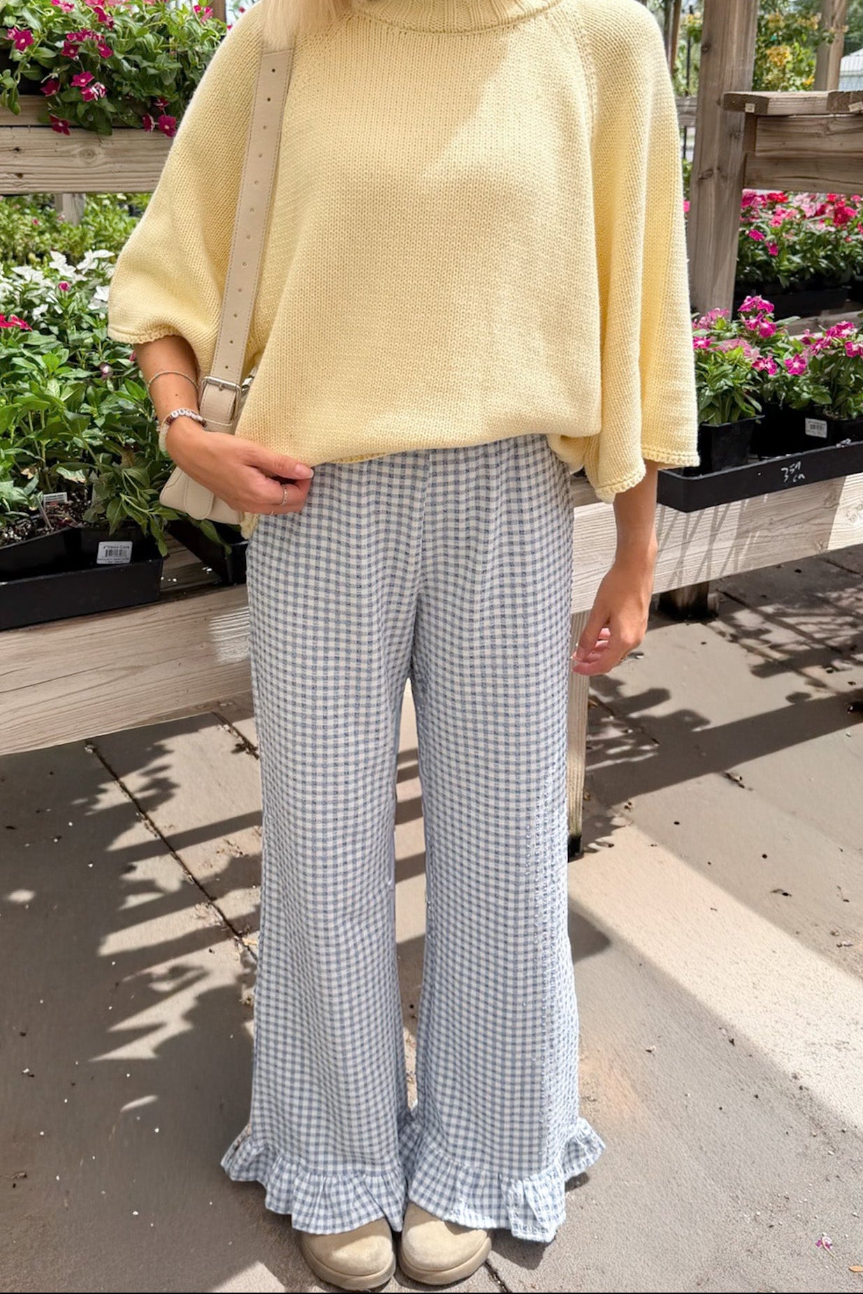 A woman wearing a pale yellow knit mock-neck tunic top and light blue and white gingham wide-leg pants with ruffled hems, standing in a garden center.