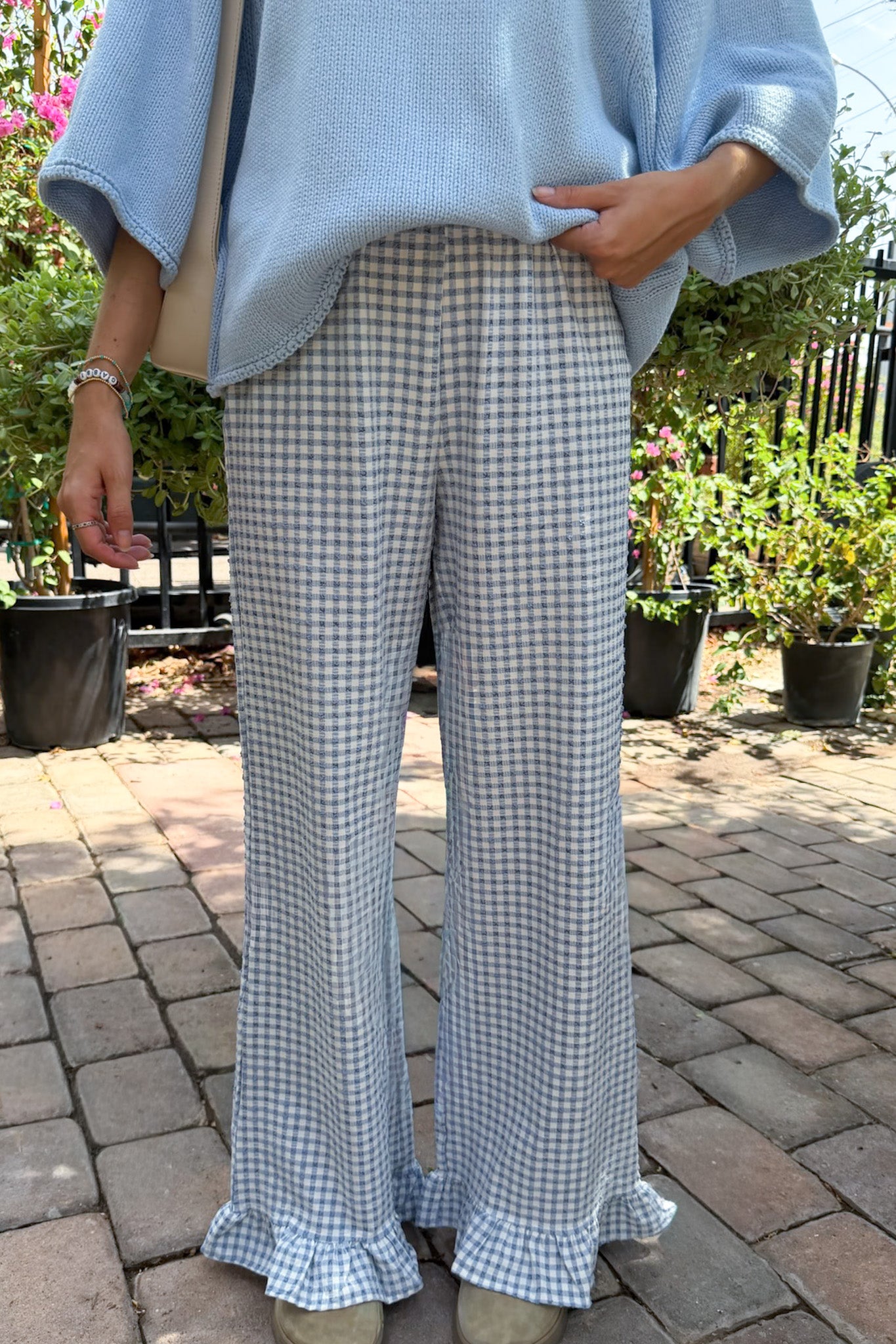 A person wearing light blue wide-leg ruffled pants with a blue and white gingham pattern, paired with a light blue knit top, standing on a brick patio with potted plants in the background.