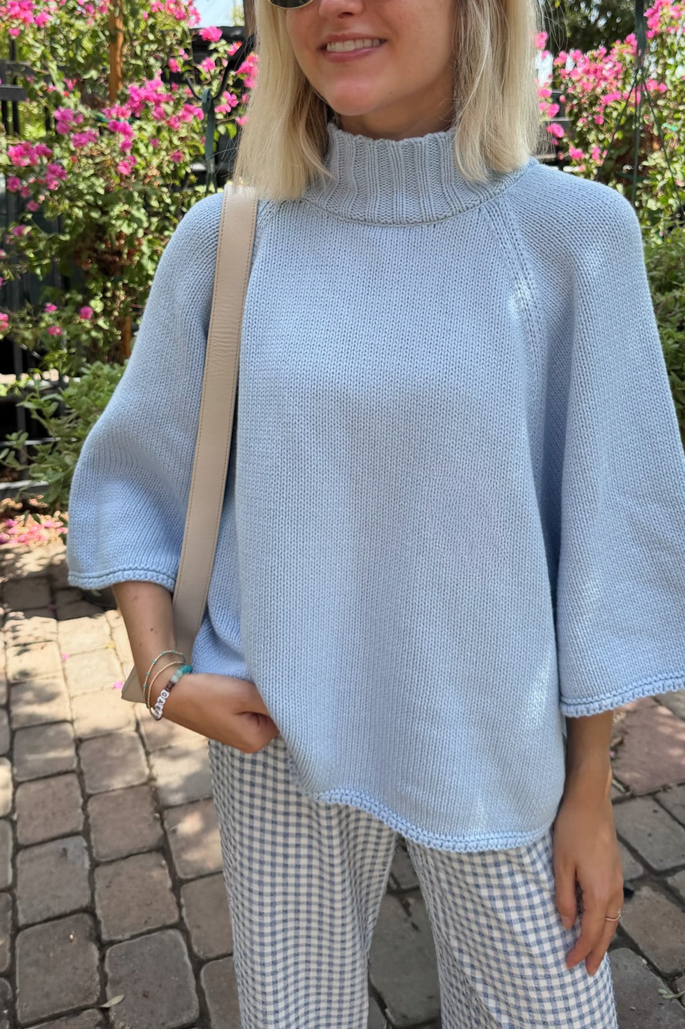 A woman wearing a light blue knit mock-neck poncho-style sweater with elbow-length sleeves, paired with gingham pants and a beige shoulder bag, standing outdoors on a brick path with pink flowers in the background.