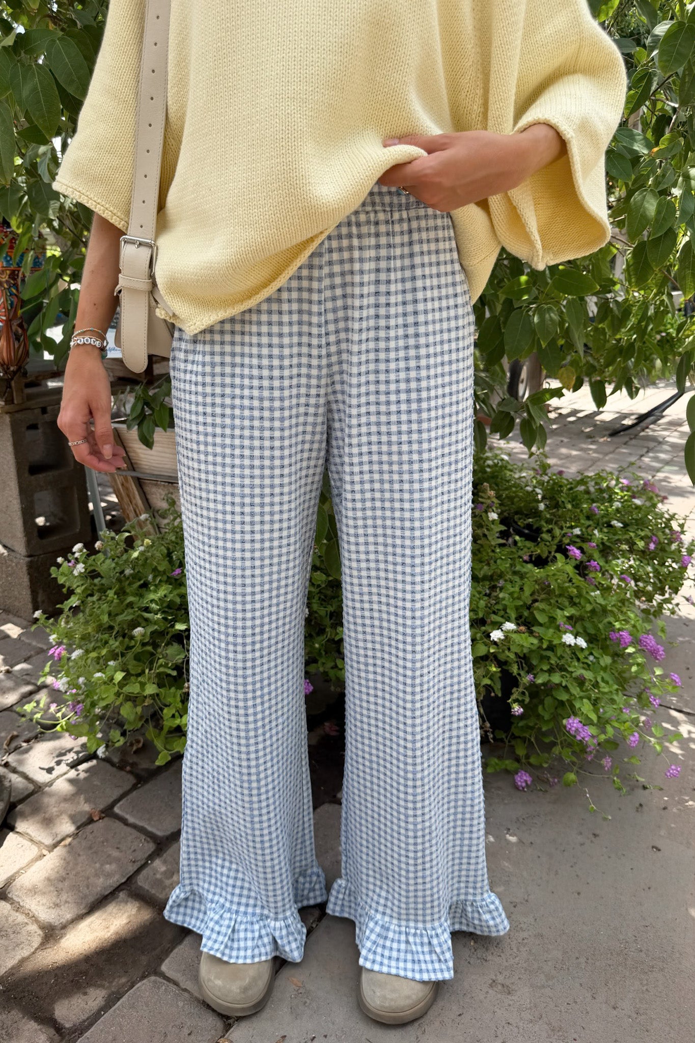 A person outdoors wearing light blue and white gingham wide-leg pants with ruffled hems, paired with a pale yellow knit top and beige shoes.