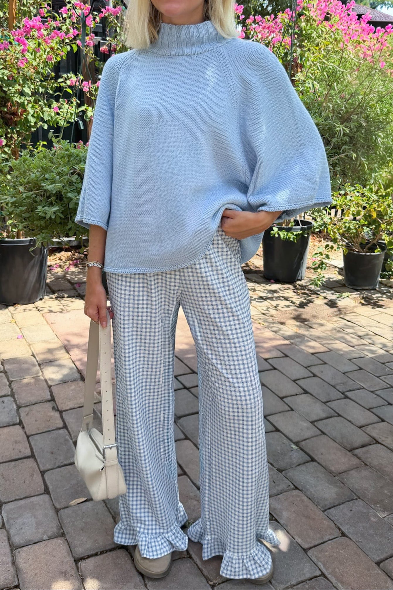 A woman wearing a light blue mock-neck knit sweater and light blue-and-white gingham wide-leg pants, standing outdoors on a brick patio with potted pink flowers in the background.