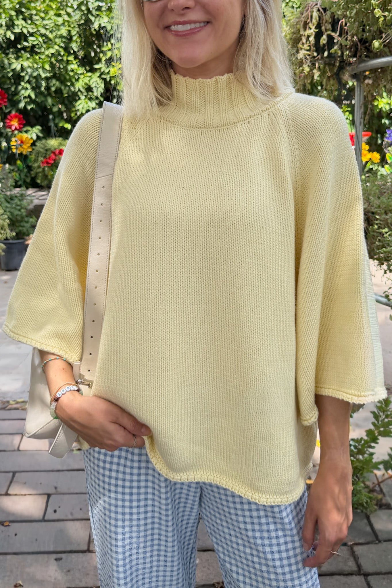 A woman wearing a pale yellow knit mock-neck tunic top with wide three-quarter sleeves, paired with blue and white gingham trousers, standing outdoors in a garden setting.