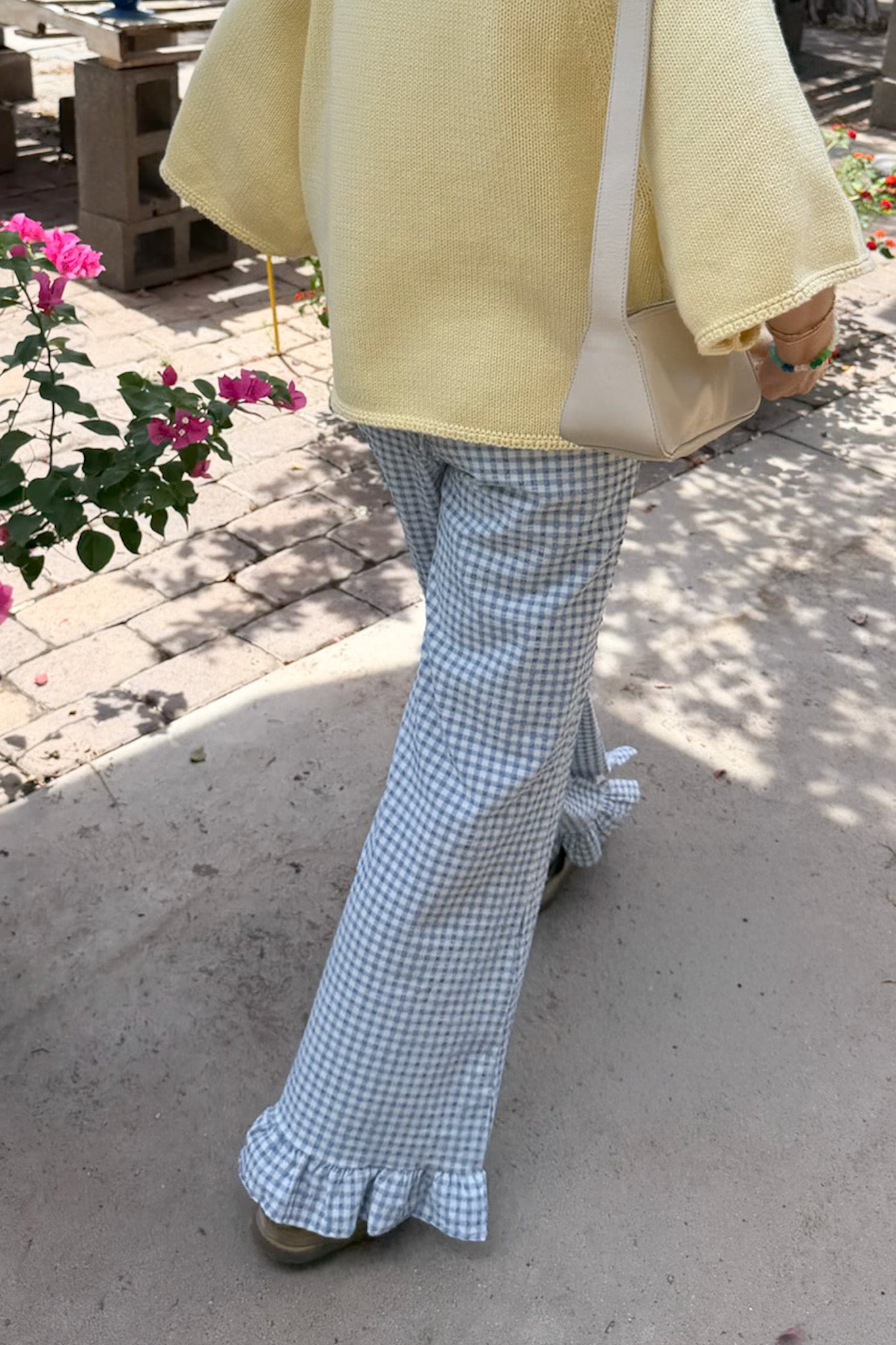 A person outdoors wearing light blue and white gingham wide-leg flare pants with ruffled hems, a pale yellow knit top, and carrying a cream shoulder bag.