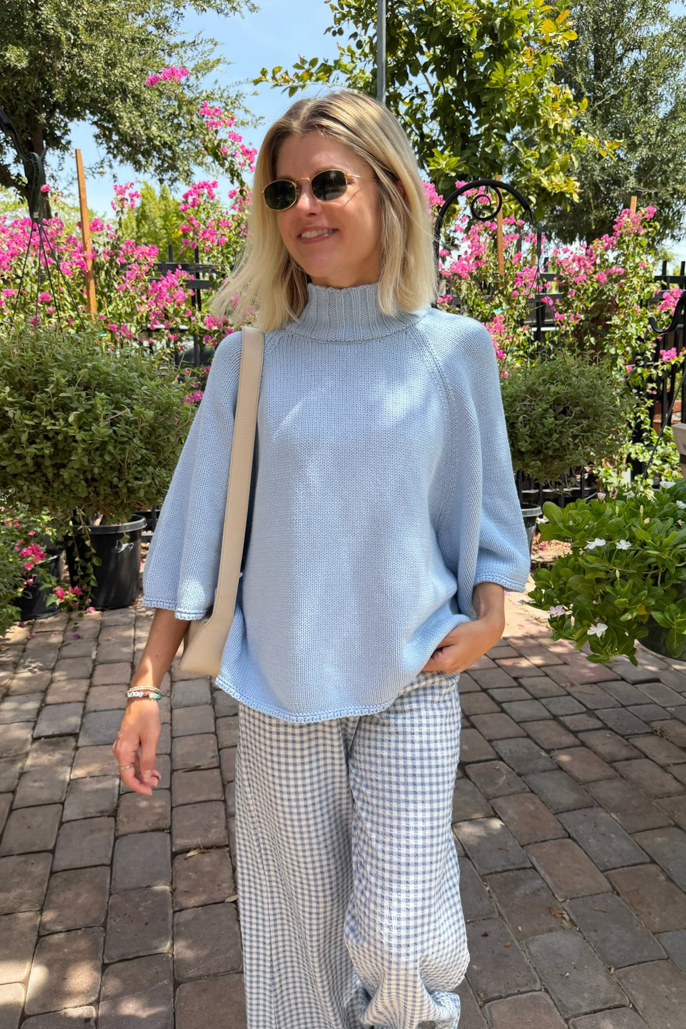 A woman wearing a light blue mock-neck knit sweater and light blue-and-white gingham wide-leg pants, standing outdoors on a brick path with pink flowers and green foliage in the background.