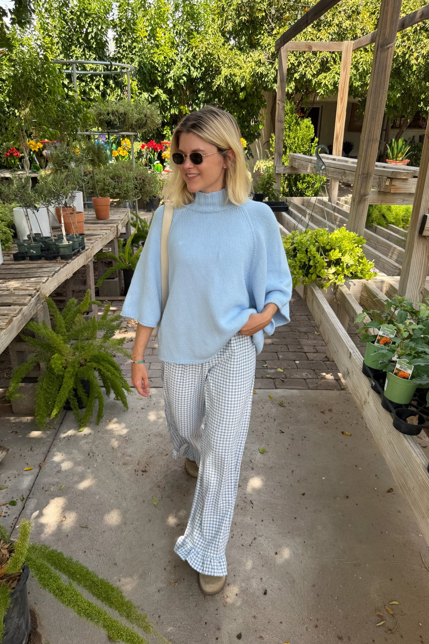 A woman wearing a light blue mock-neck tunic top and white-and-blue gingham wide-leg pants, walking in a sunny garden center.