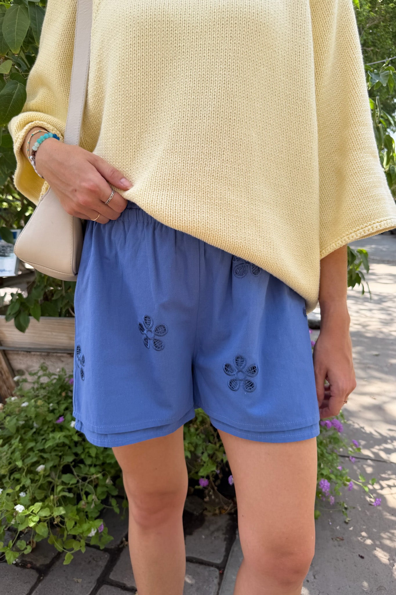 A person outdoors wearing blue shorts with embroidered floral details and a pale yellow knit top, with a light-colored shoulder bag visible.