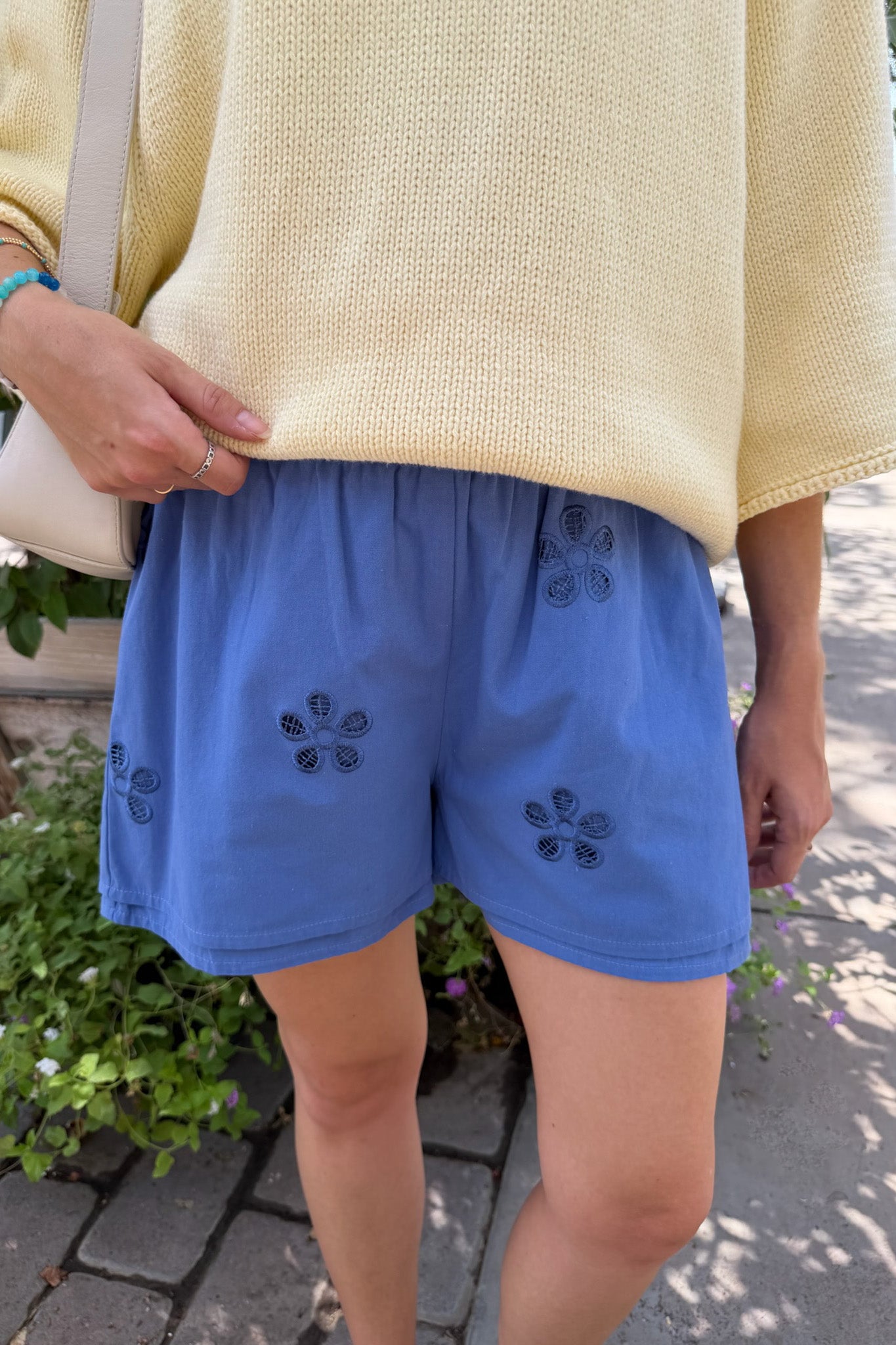 A person outdoors wearing blue shorts with dark embroidered floral motifs and a pale yellow knit top, with a light-colored shoulder bag partially visible.