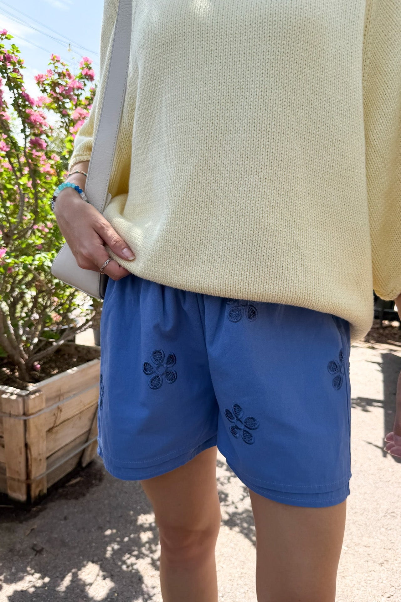 A person outdoors wearing blue shorts with dark floral embroidery and a pale yellow knit top, standing near a planter with pink flowers.
