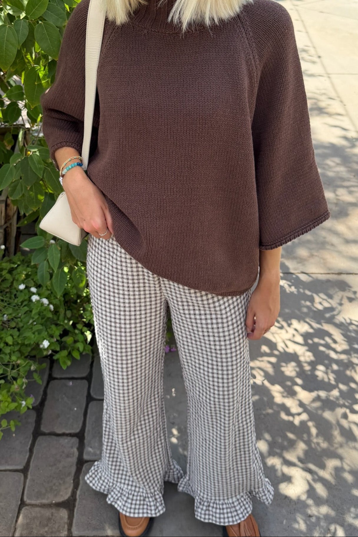 A woman wearing a brown ribbed knit mock-neck tunic top with elbow-length sleeves, paired with black and white gingham wide-leg pants with ruffled hems, standing outdoors on a paved walkway.