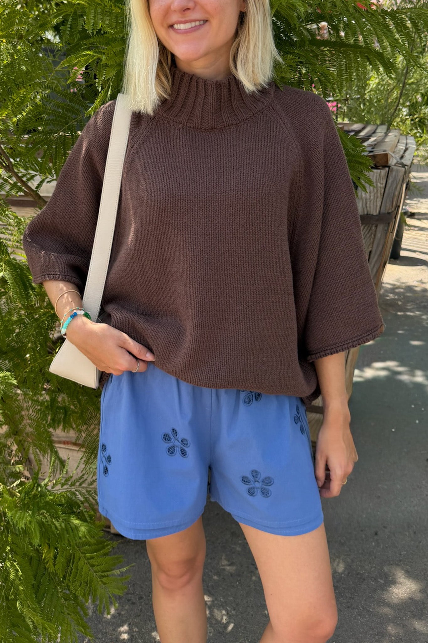 A woman wearing a brown mock-neck knit top and blue shorts with dark floral embroidery, standing outdoors next to green foliage.