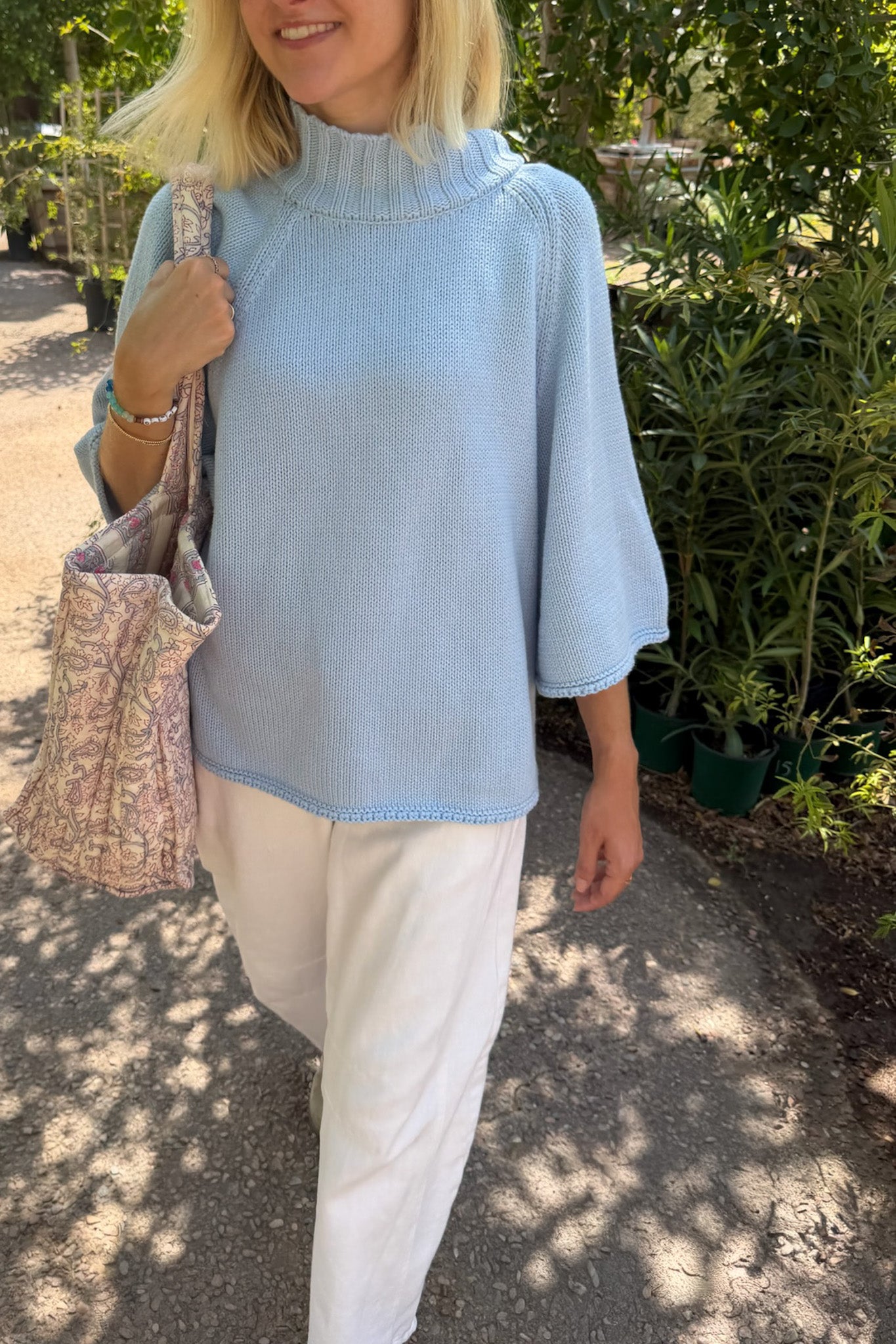 A woman outdoors wearing a light blue knit mock-neck tunic top with elbow-length sleeves and white trousers, carrying a floral patterned tote bag.