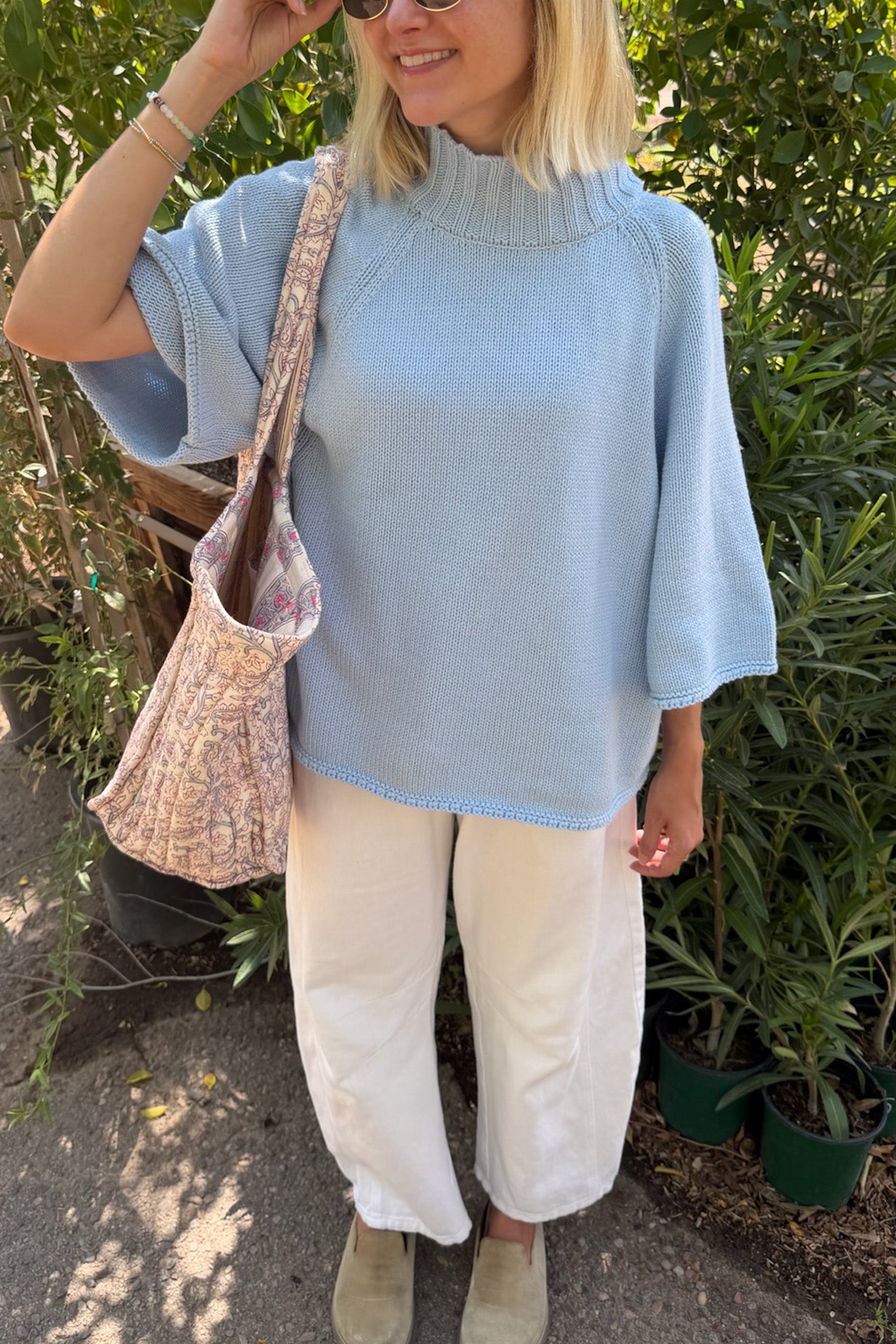 A woman wearing a light blue knit mock-neck tunic sweater with wide three-quarter sleeves, paired with cream wide-leg trousers and beige shoes, carrying a floral patterned shoulder bag in an outdoor garden setting.