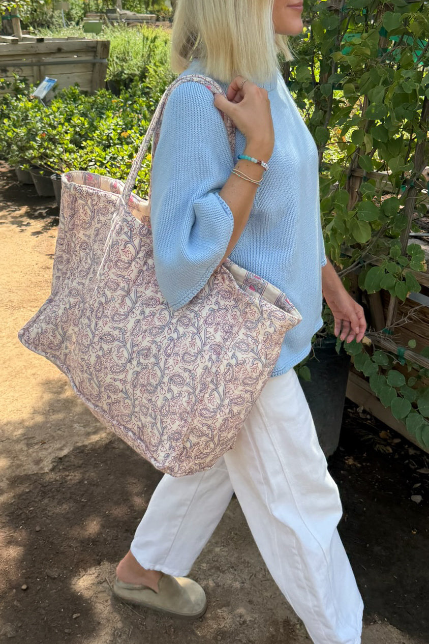A woman in a light blue sweater and white pants carries a large patterned tote bag with a pink, beige, and white paisley design in an outdoor garden setting.