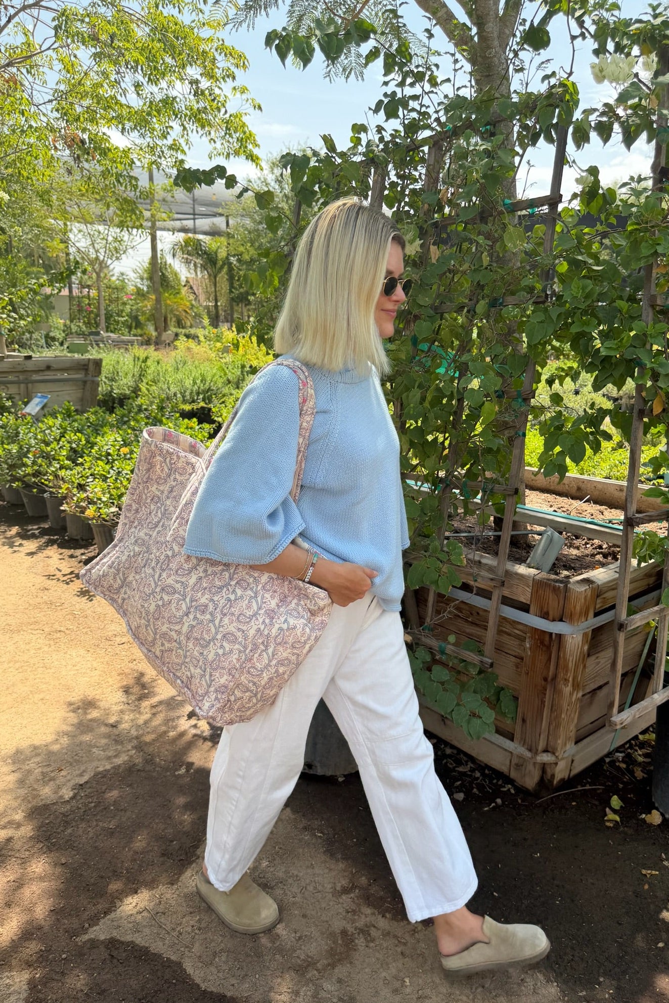 A woman walking outdoors in a garden wearing a light blue sweater, white pants, beige shoes, and carrying a large pink floral tote bag.
