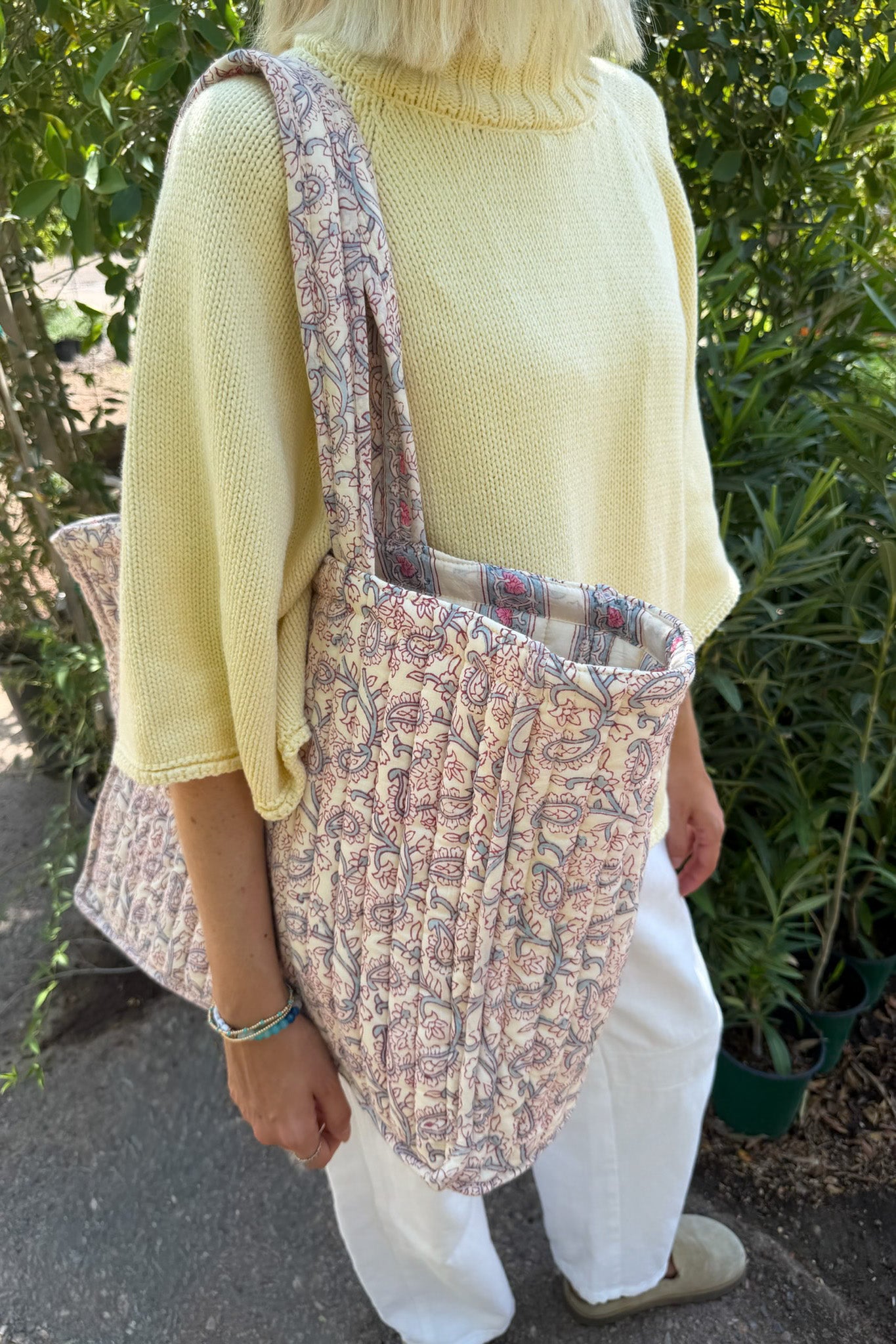 A person outdoors wearing a pale yellow knit sweater and white pants, carrying a large quilted fabric tote bag with a floral and paisley pattern in muted pink, beige, and blue tones.