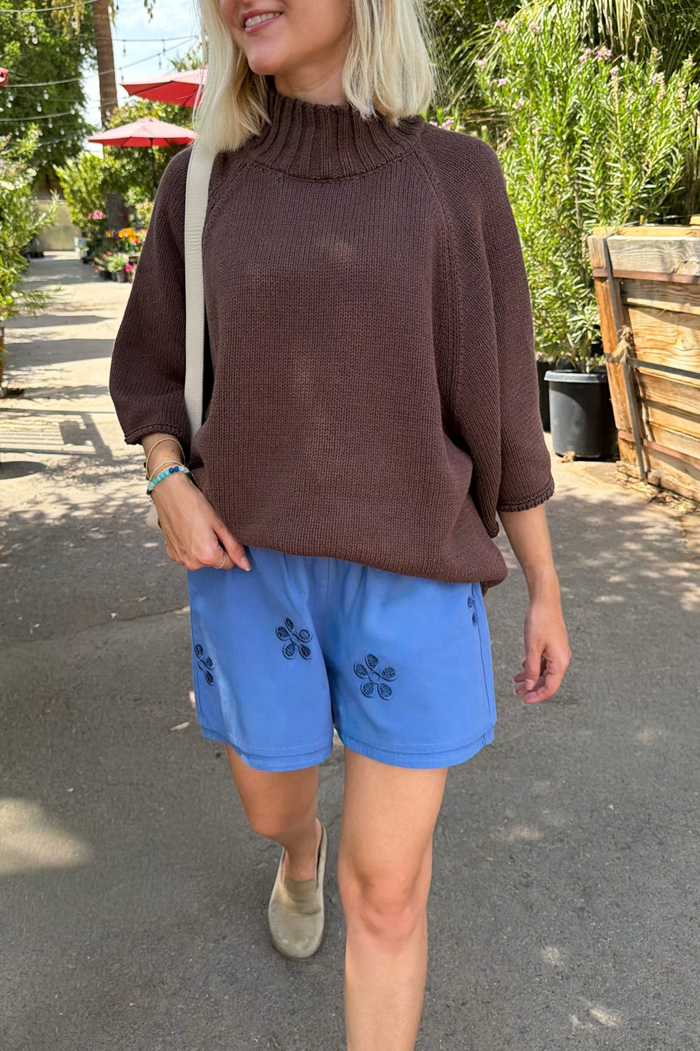 A woman wearing a brown ribbed mock-neck sweater and blue shorts with black floral embroidery, walking outdoors on a paved path with greenery in the background.