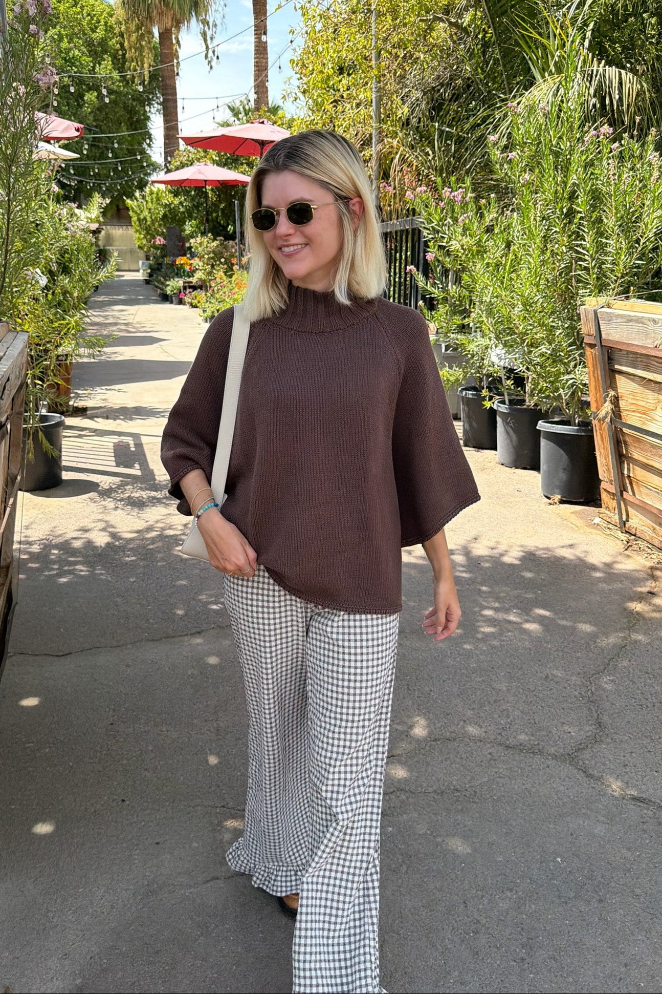 A woman walking outdoors on a paved path wearing a brown mock-neck short-sleeve top and black-and-white gingham wide-leg pants, with a white shoulder bag. 