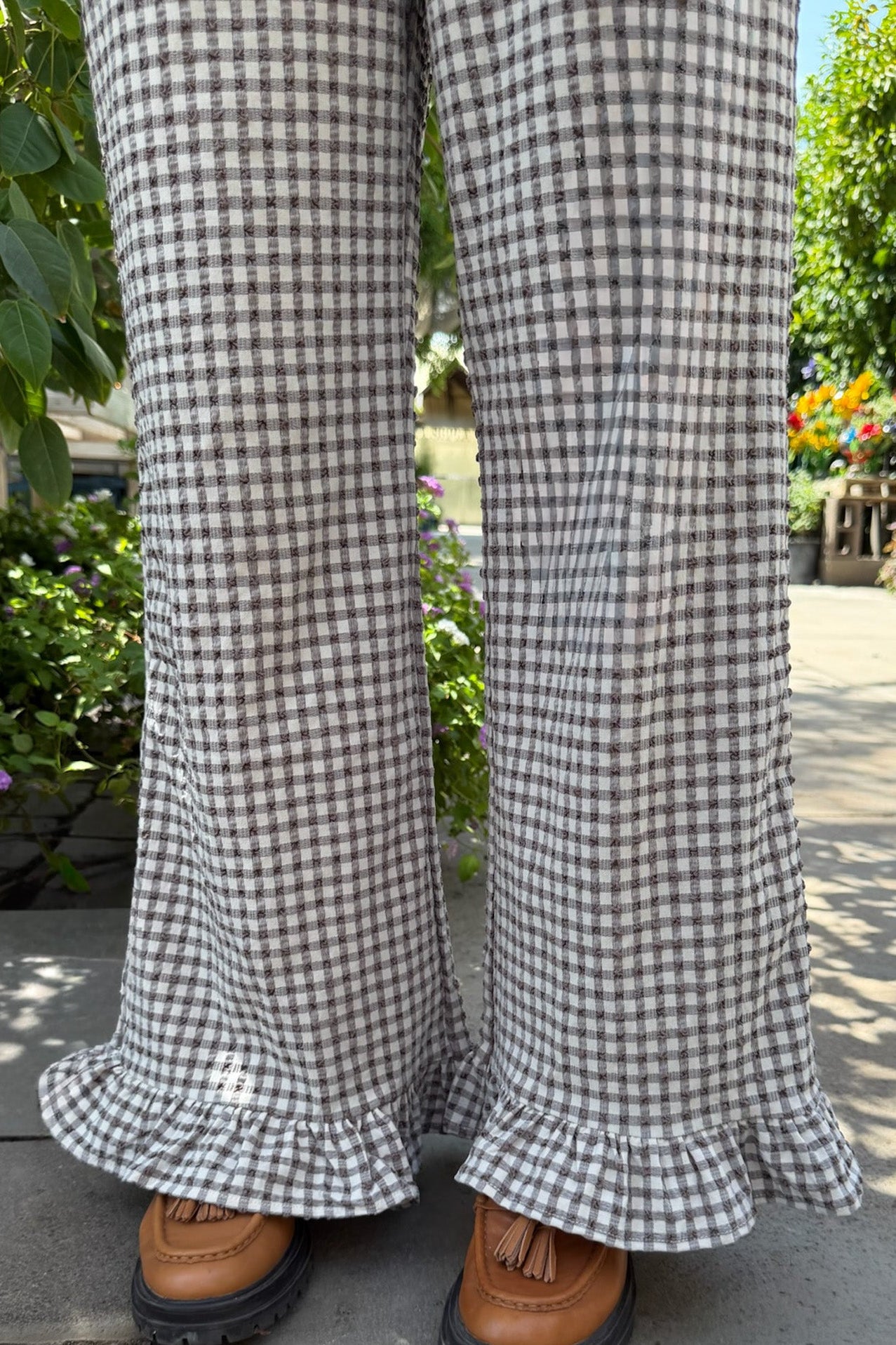 A close-up of a person's legs wearing wide-leg gingham plaid pants with ruffled hems, paired with brown leather tassel loafers, standing on a paved path in a garden setting. 