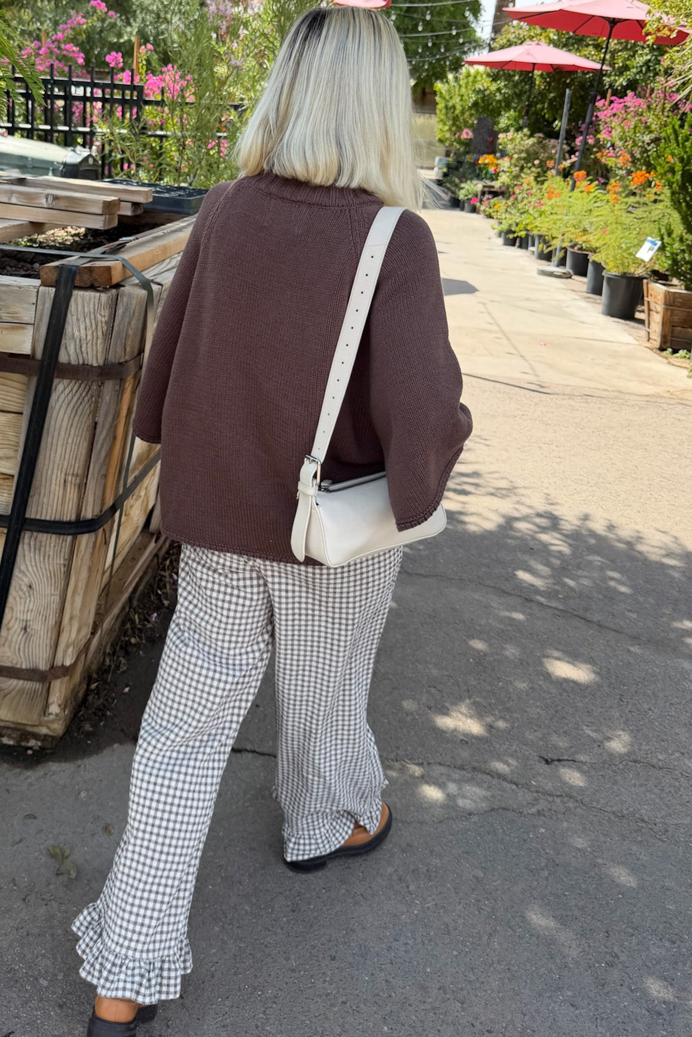 A woman viewed from behind wearing a brown knit sweater, black and white gingham wide-leg pants with ruffled hems, and carrying a white shoulder bag with a black strap, walking along a garden path.