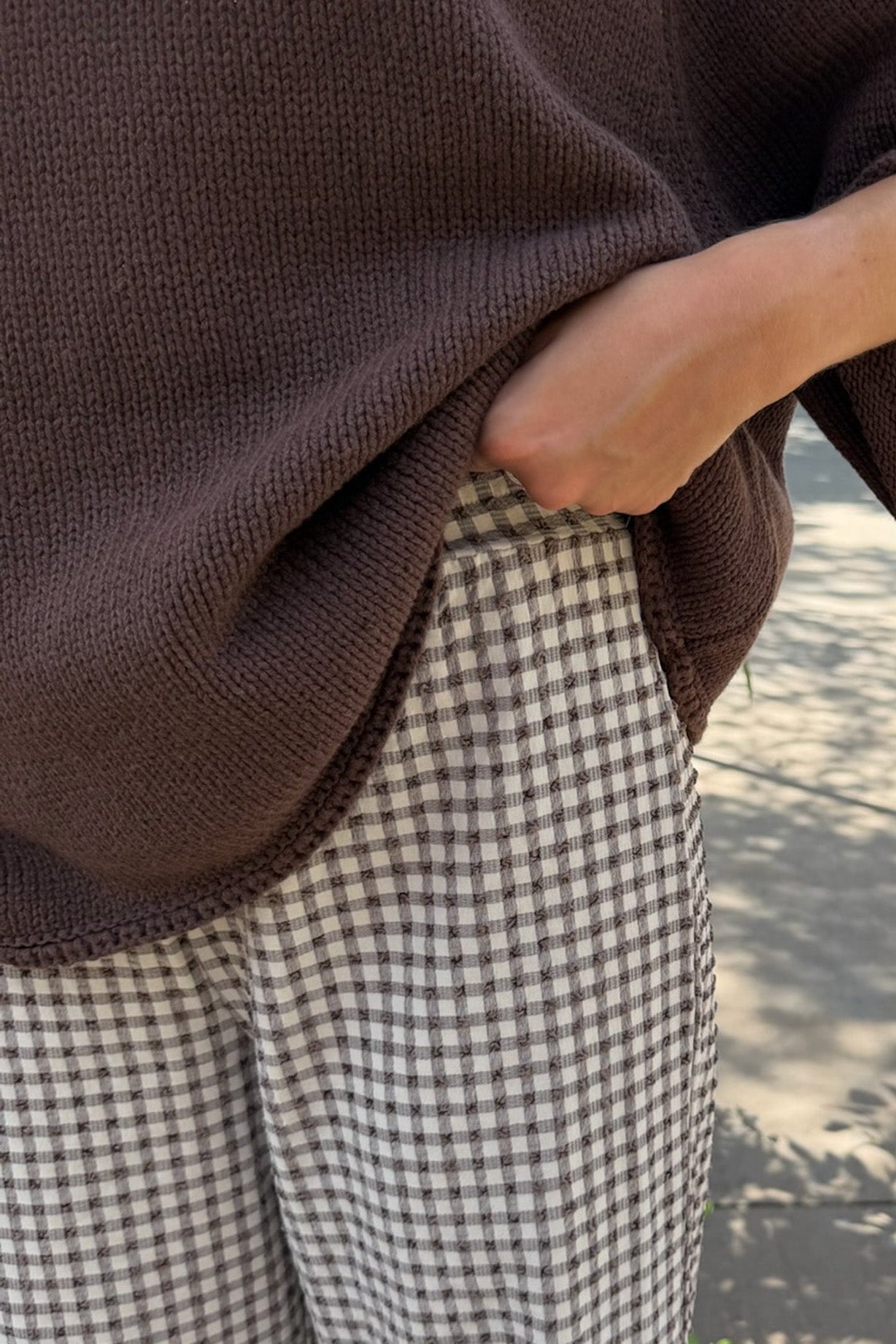 A close-up lifestyle shot of a person wearing a dark brown ribbed knit sweater and brown-and-white gingham checkered pants, with their hand pulling the hem of the sweater down.
