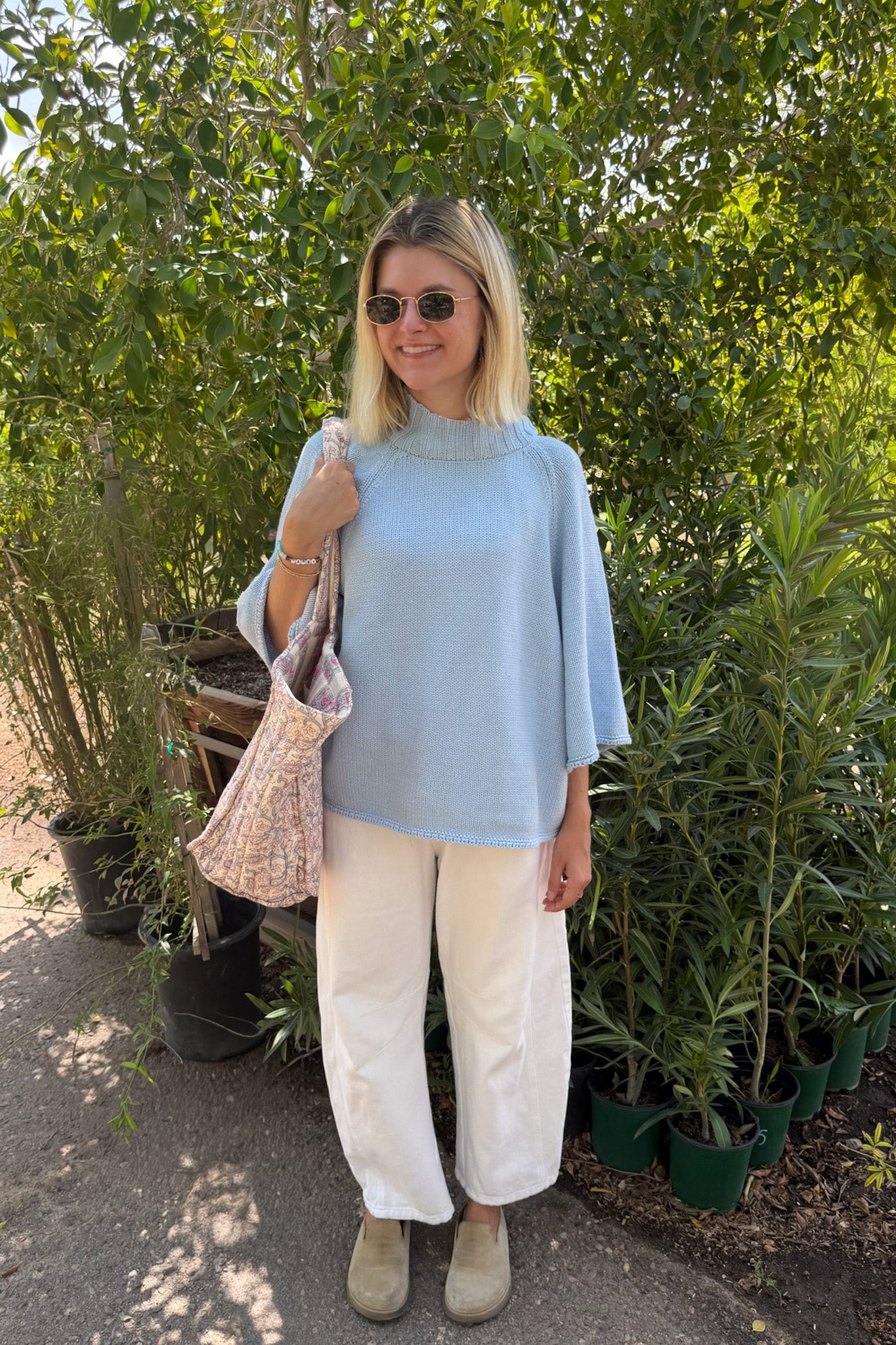 A woman wearing a light blue mock-neck top, white wide-leg pants, and beige shoes stands outdoors in a garden setting, holding a pink patterned tote bag.