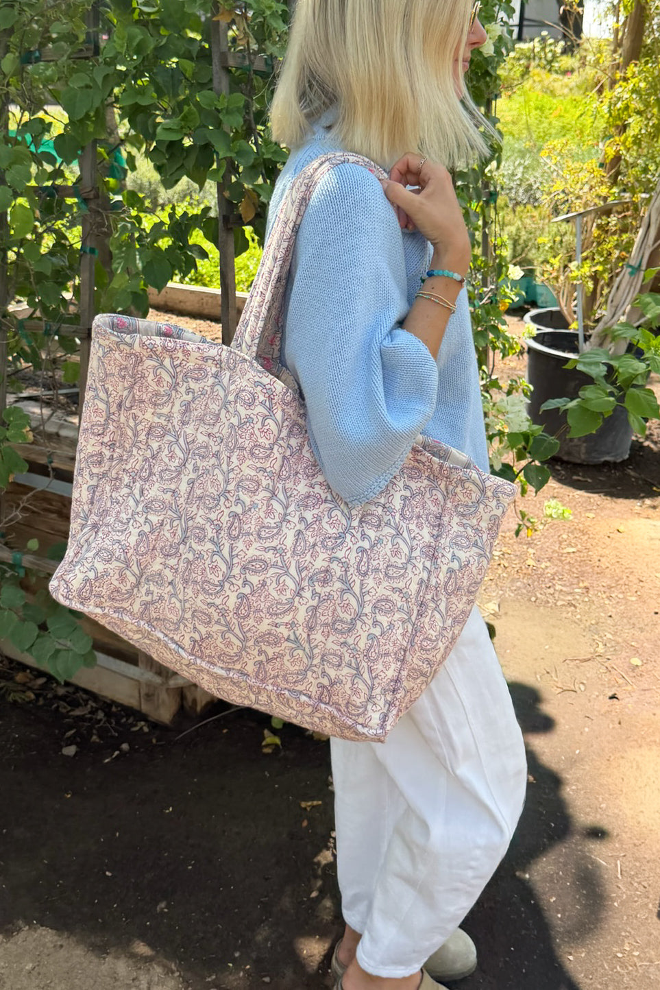 A woman in a blue sweater and white pants carries a large patterned tote bag with pink, beige, and blue floral motifs in an outdoor garden setting.
