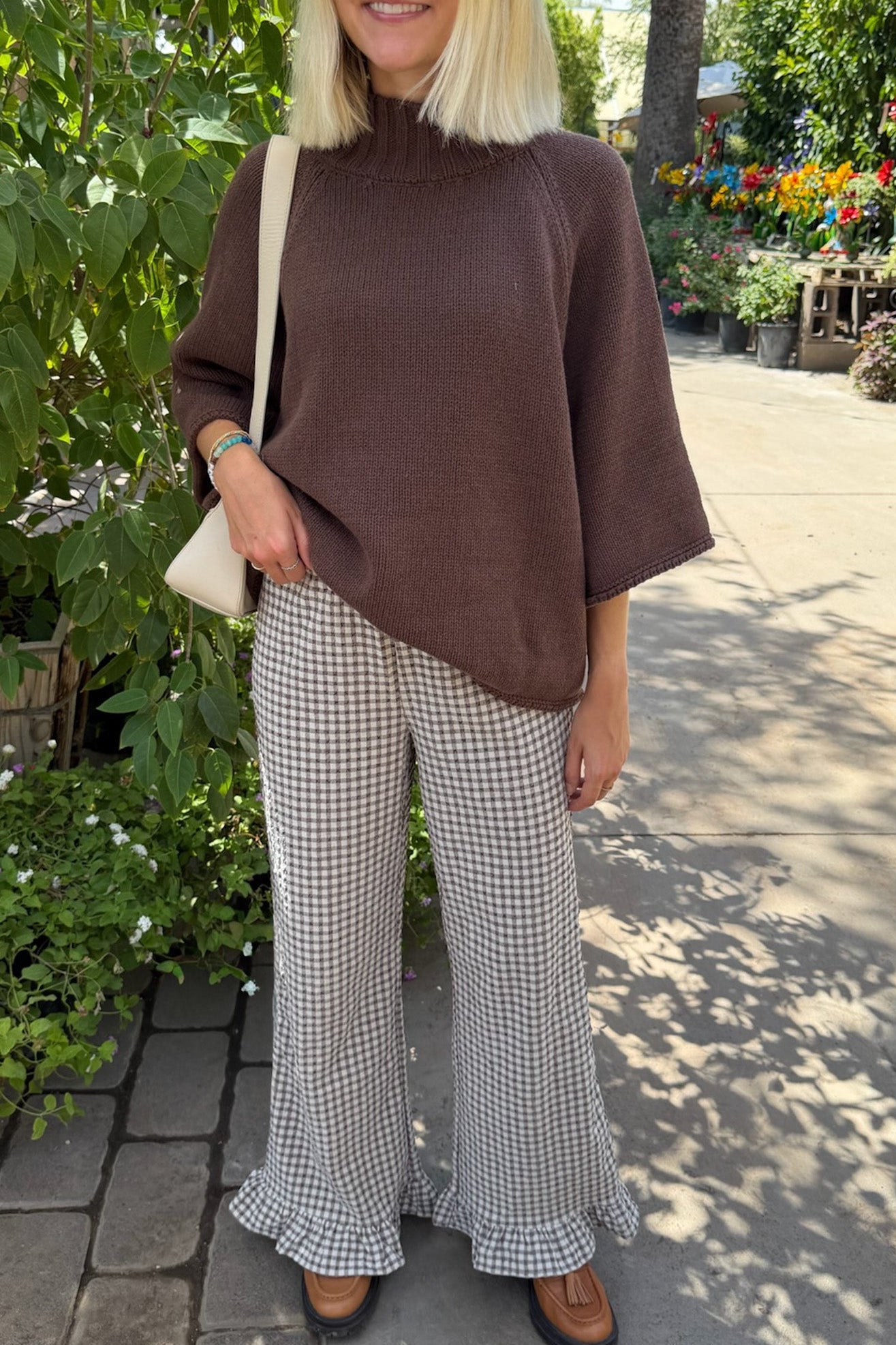 A woman wearing a brown mock-neck knit sweater and black-and-white gingham wide-leg flared pants with ruffled hems, standing outdoors in a garden setting.