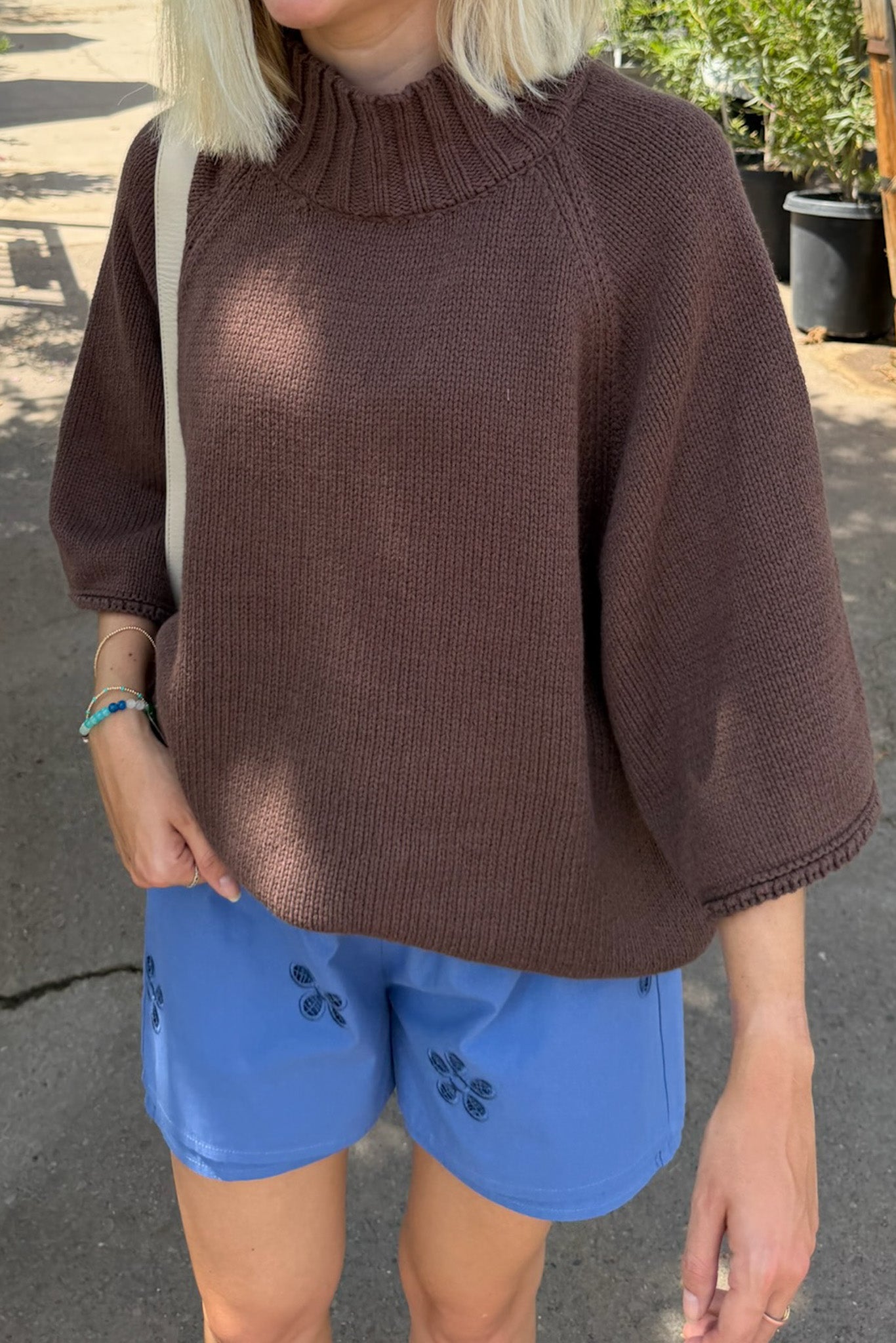 A woman wearing a brown ribbed knit mock-neck sweater with elbow-length raglan sleeves, paired with blue shorts, standing outdoors.