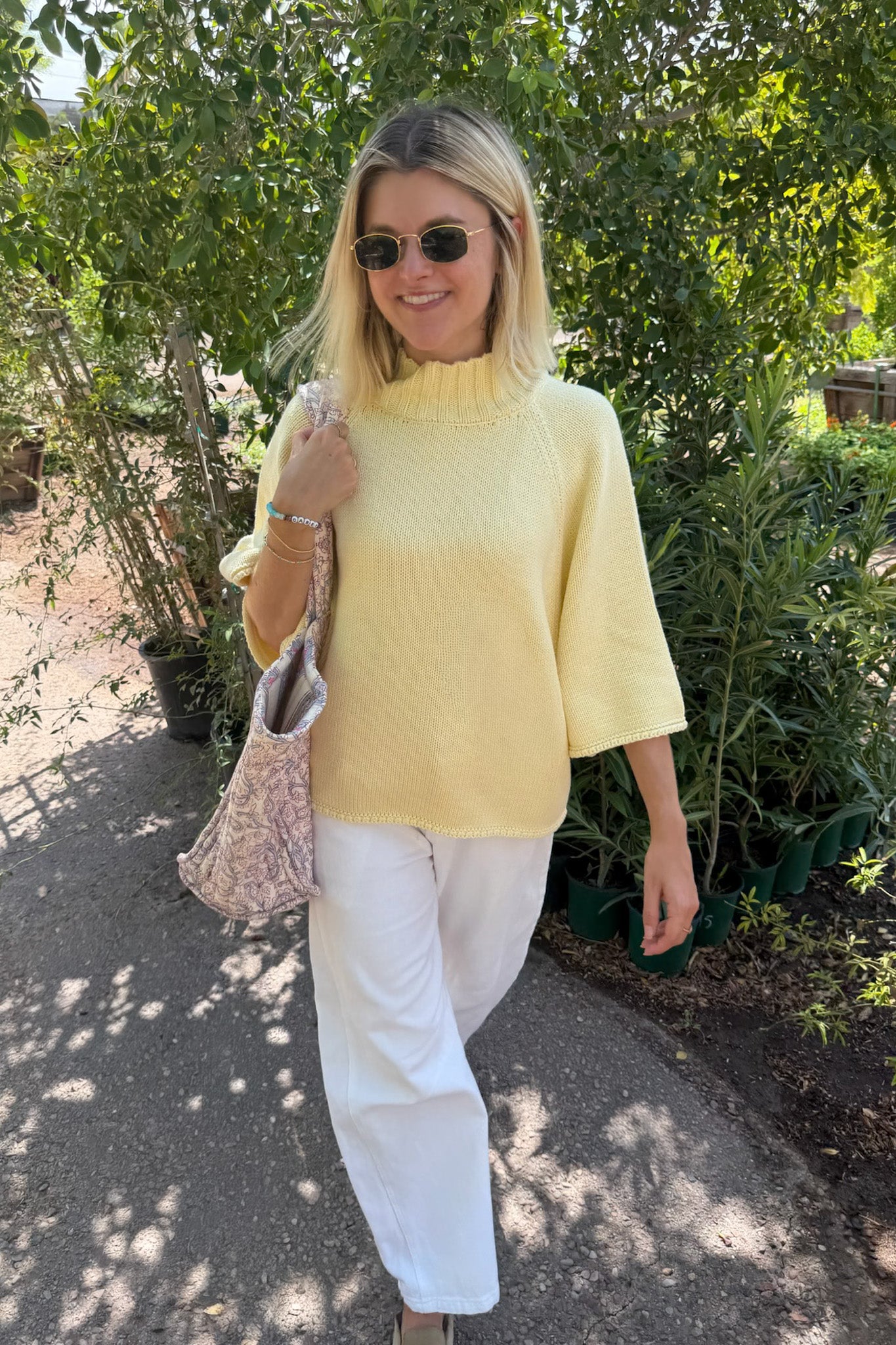 A woman wearing a pale yellow knit mock-neck top and white trousers walks outdoors on a paved path surrounded by green plants. She is holding a patterned shoulder bag.