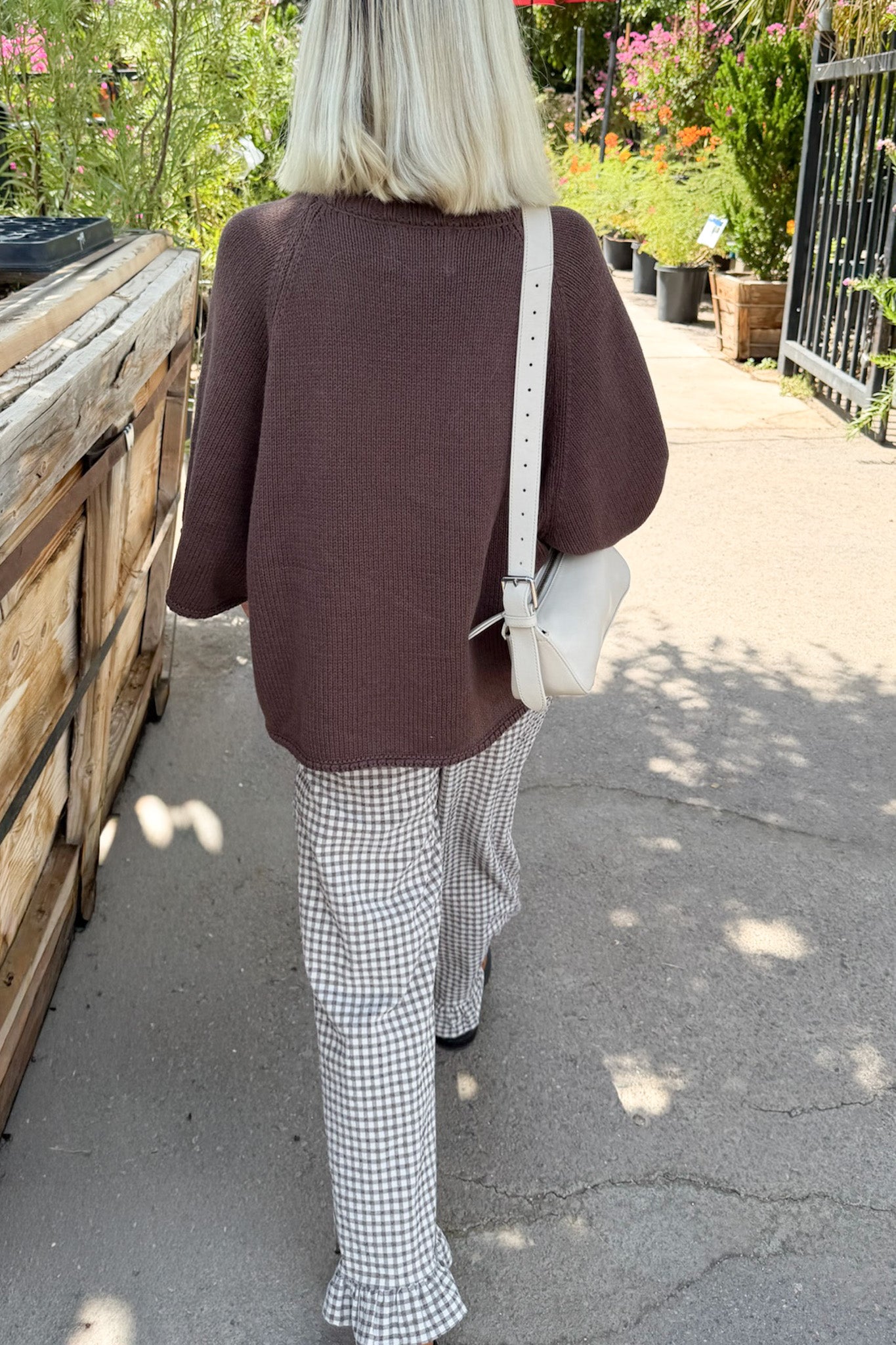 A woman viewed from behind wearing a brown knit sweater and white-and-black gingham flared pants, carrying a white shoulder bag in an outdoor garden walkway.