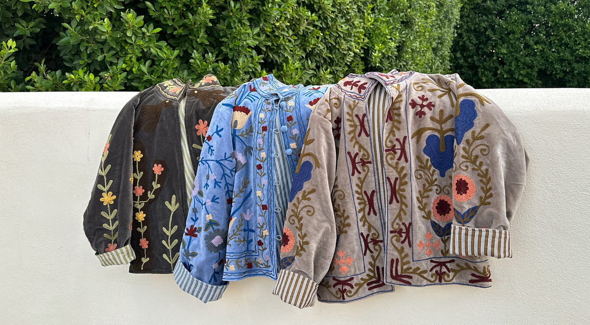 Four embroidered bomber jackets in black, blue, and beige tones draped over a white wall with green hedges in the background.