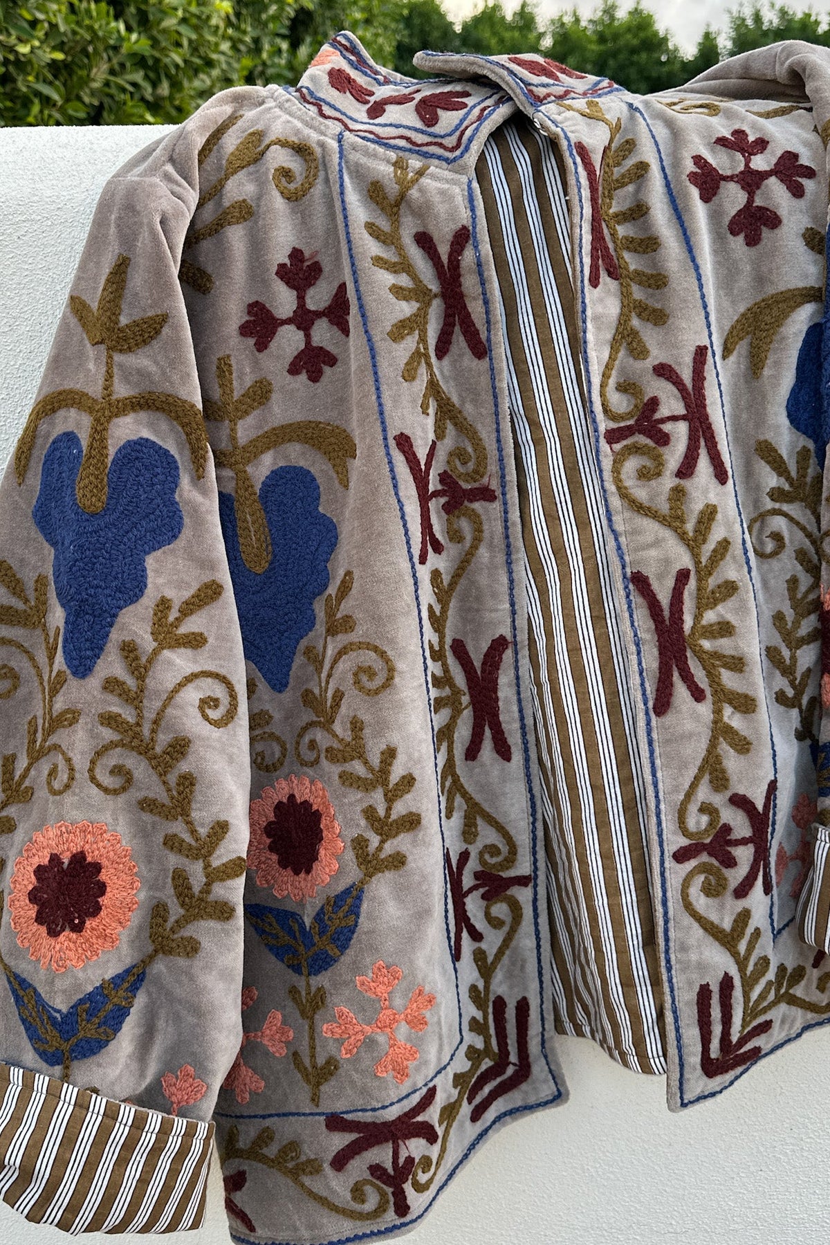 A beige embroidered cardigan or jacket with blue, burgundy, and gold floral patterns, draped outdoors on a white surface with green foliage in the background.