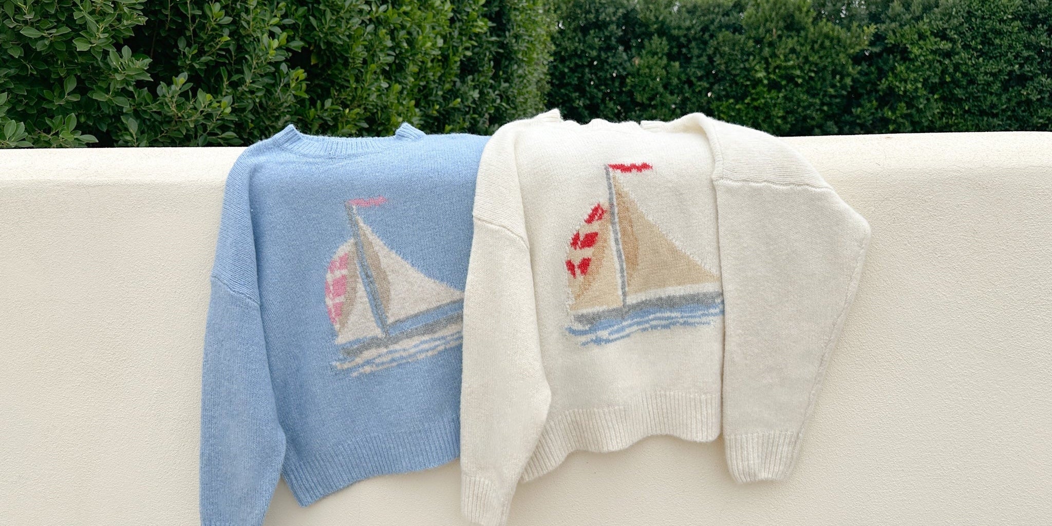 Two sweaters with sailboat designs hanging on a railing outdoors.