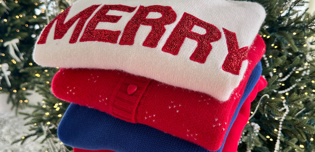 Stack of colorful sweaters with 'MERRY' text in front of a Christmas tree.