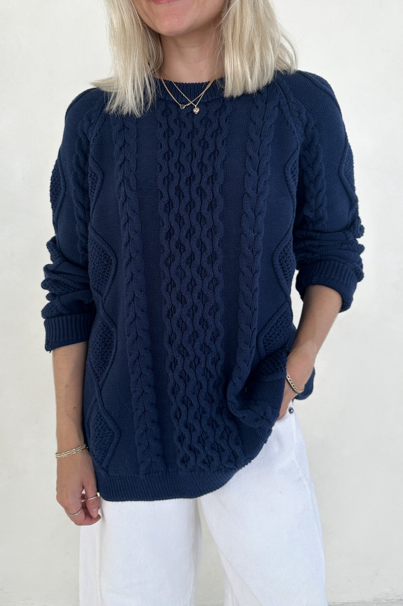 Person wearing a navy blue cable knit sweater and white pants against a plain background