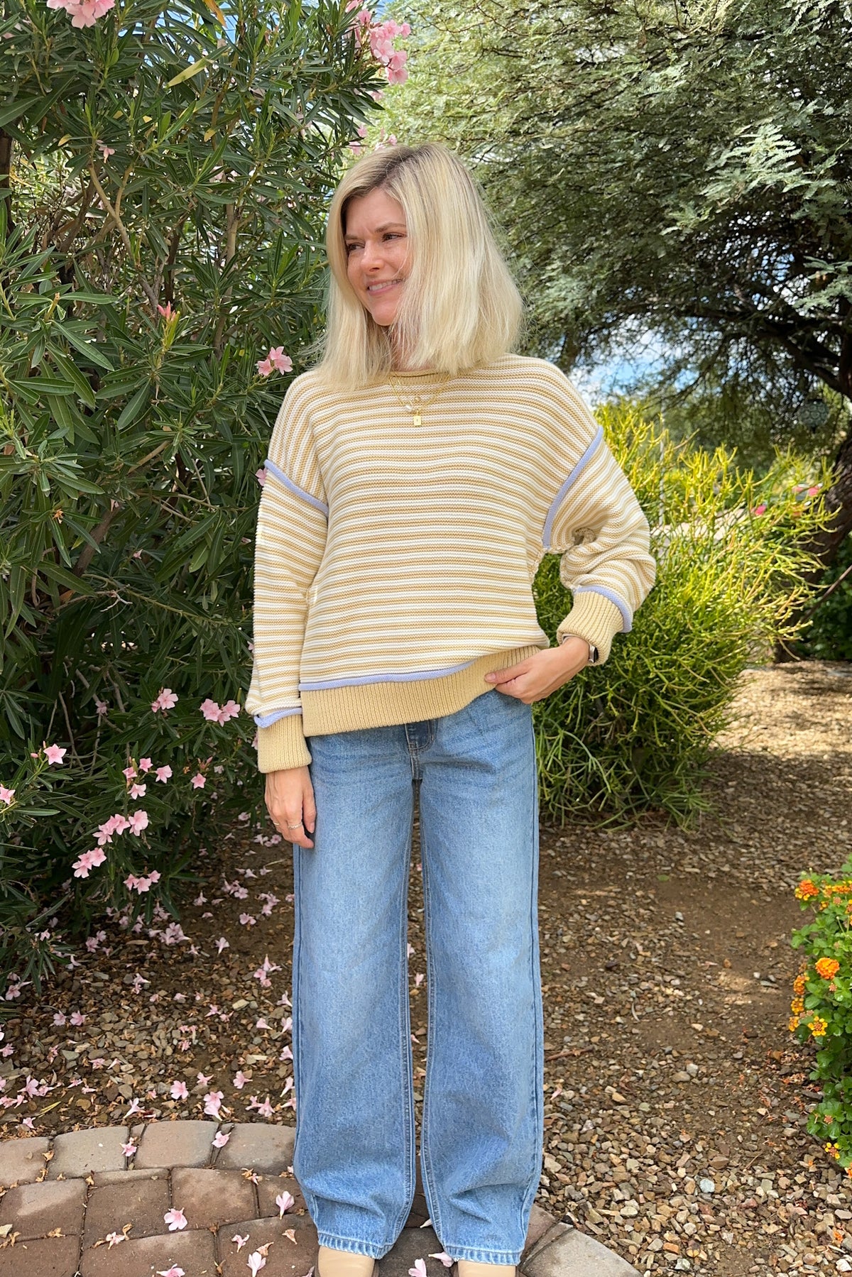 A woman with blonde hair stands outdoors in a garden setting, wearing a beige and white striped long-sleeve knit sweater with light blue trim, paired with wide-leg light blue jeans.