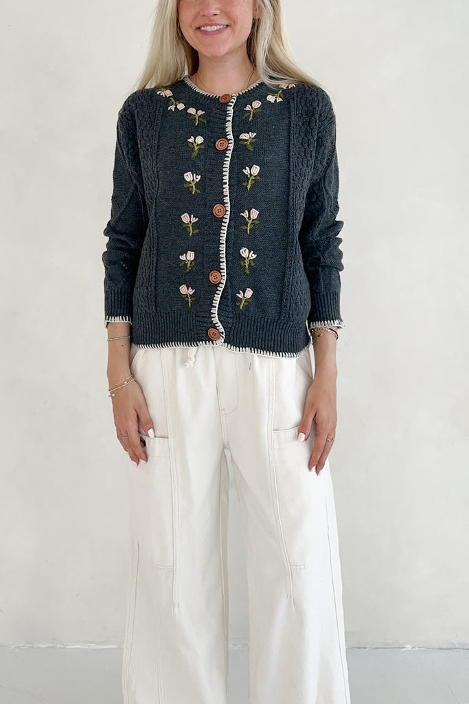 Woman wearing a dark blue cardigan with floral patterns and white pants on a plain background