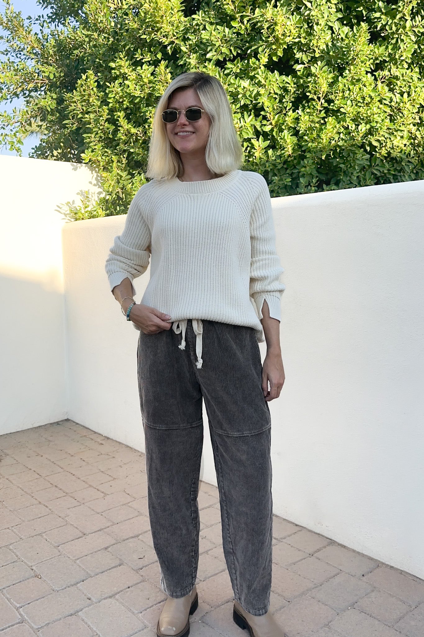 A woman wearing a cream ribbed knit sweater and dark gray drawstring sweatpants, standing outdoors against a white wall and green trees.