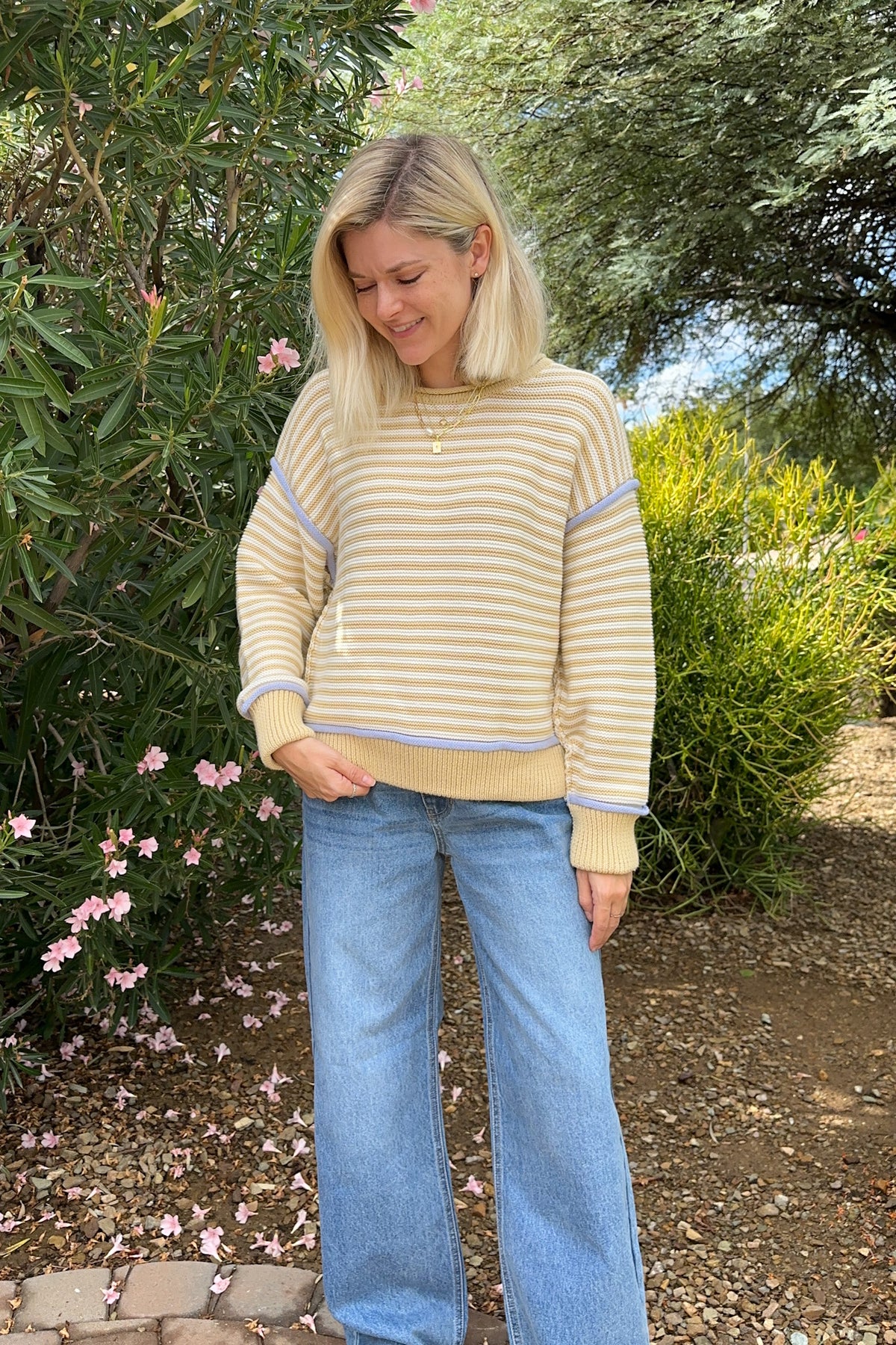 A woman wearing a beige and white striped long-sleeve sweater with light blue wide-leg jeans, standing outdoors in a garden setting.