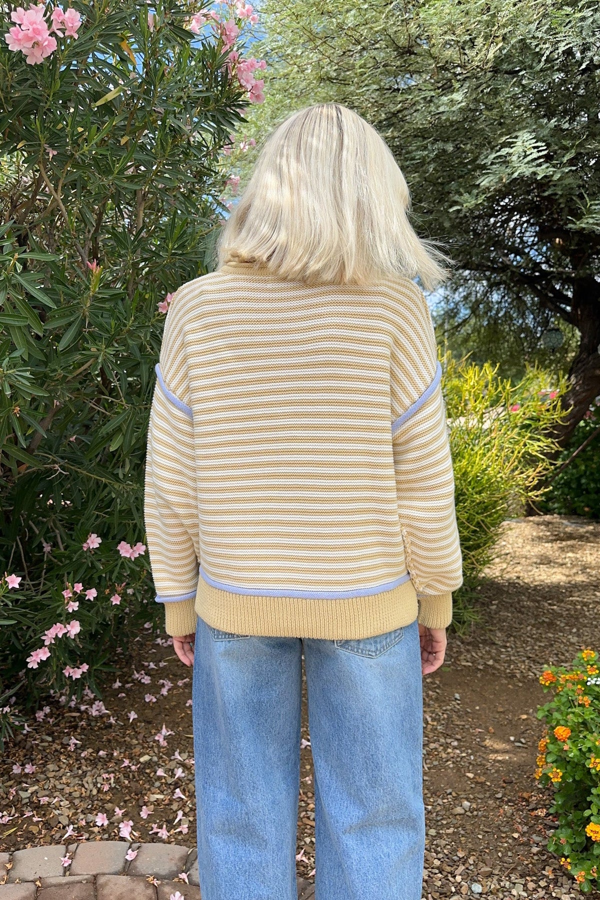 A person standing outdoors in a garden, viewed from behind, wearing a cream and tan striped knit sweater with light blue contrast panels on the shoulders and hem, paired with light blue jeans.