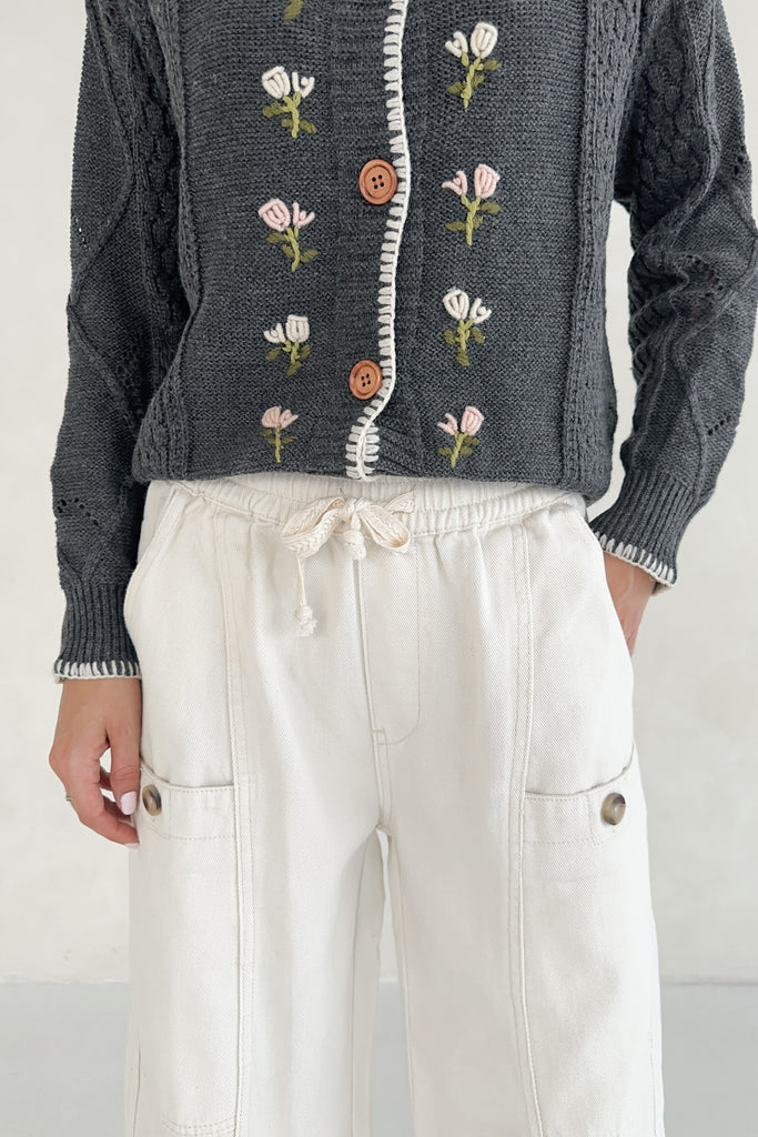 Person wearing a dark gray cardigan with floral embroidery and white pants on a plain background