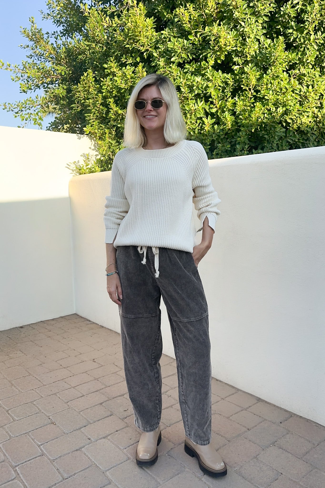 A woman wearing a cream ribbed knit sweater, dark gray corduroy pants, and beige ankle boots, standing outdoors against a white wall and green foliage.