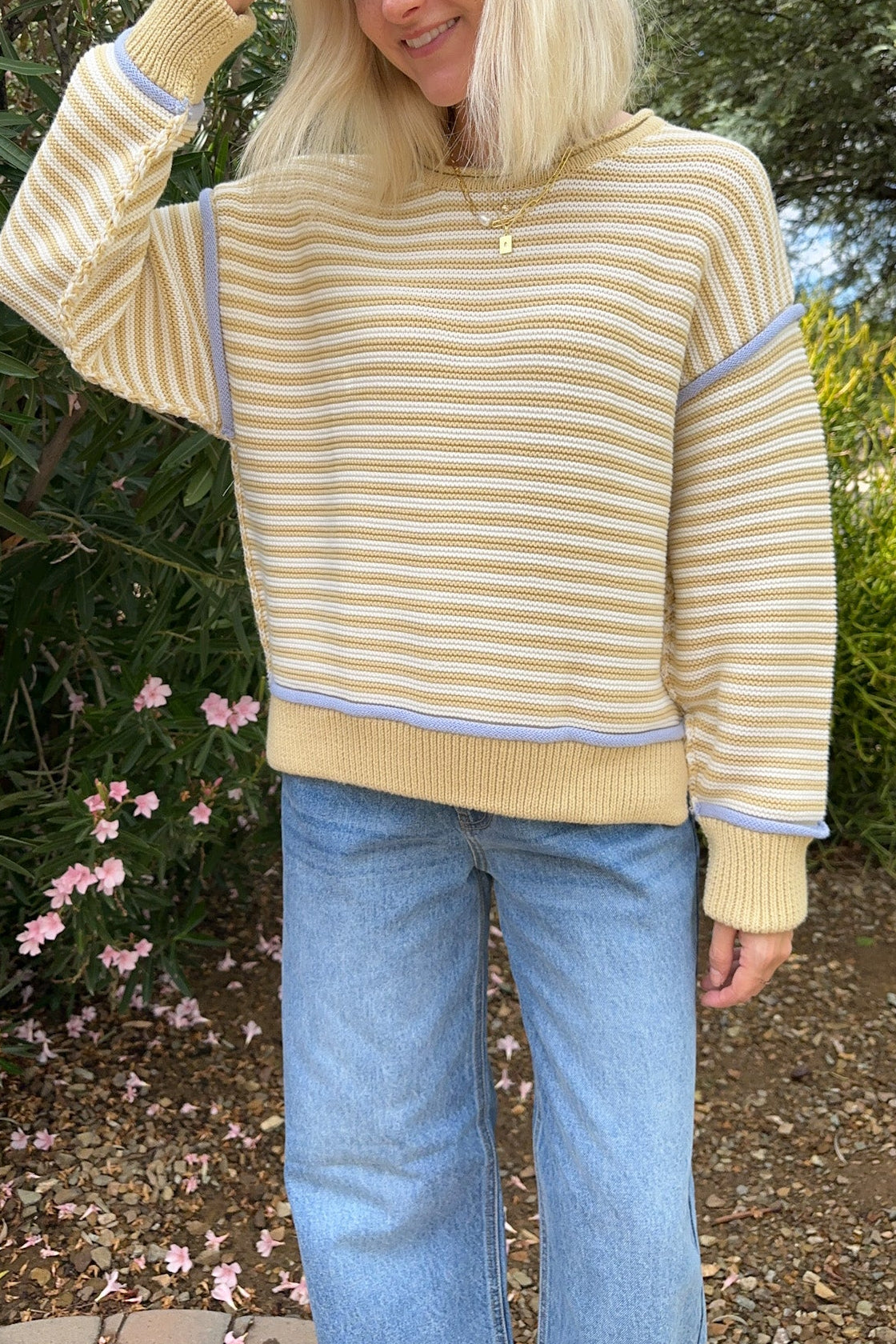 A woman wearing a yellow and beige striped knit sweater with light blue jeans, standing outdoors in a garden setting.