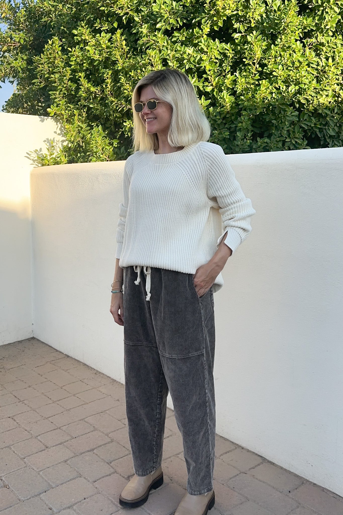 A woman wearing a cream ribbed knit sweater and dark gray corduroy pants, standing outdoors against a white wall and green foliage.