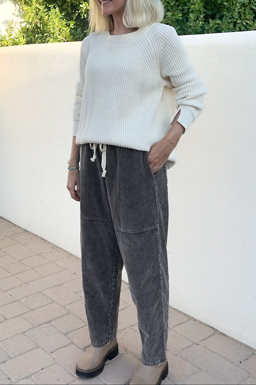 A woman with blonde hair and sunglasses stands outdoors wearing a cream ribbed knit sweater and dark gray corduroy pants, posing in front of a white wall and green foliage.