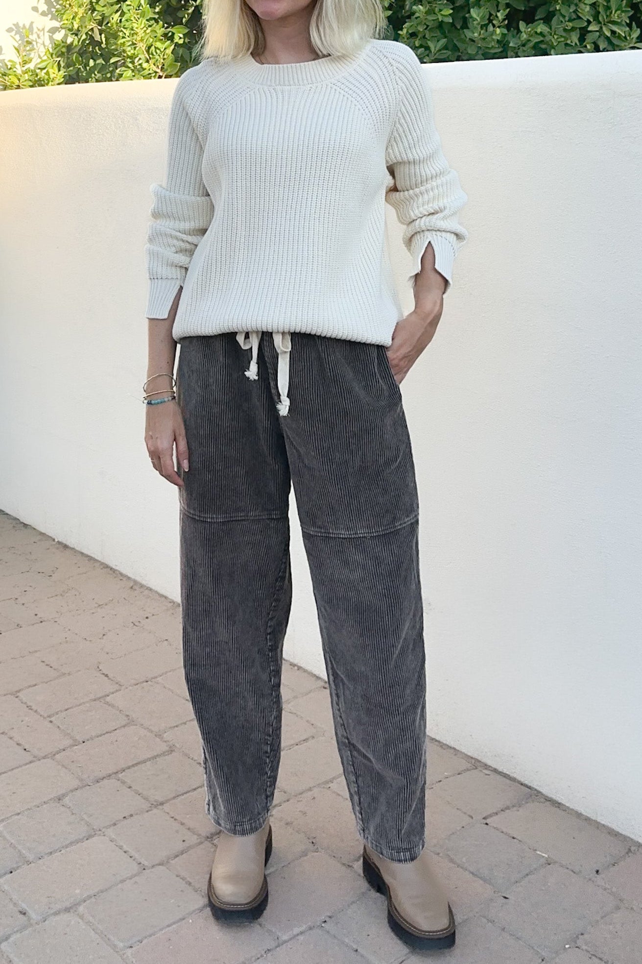 A woman wearing a cream ribbed knit sweater and dark gray corduroy pants, standing outdoors against a white wall.
