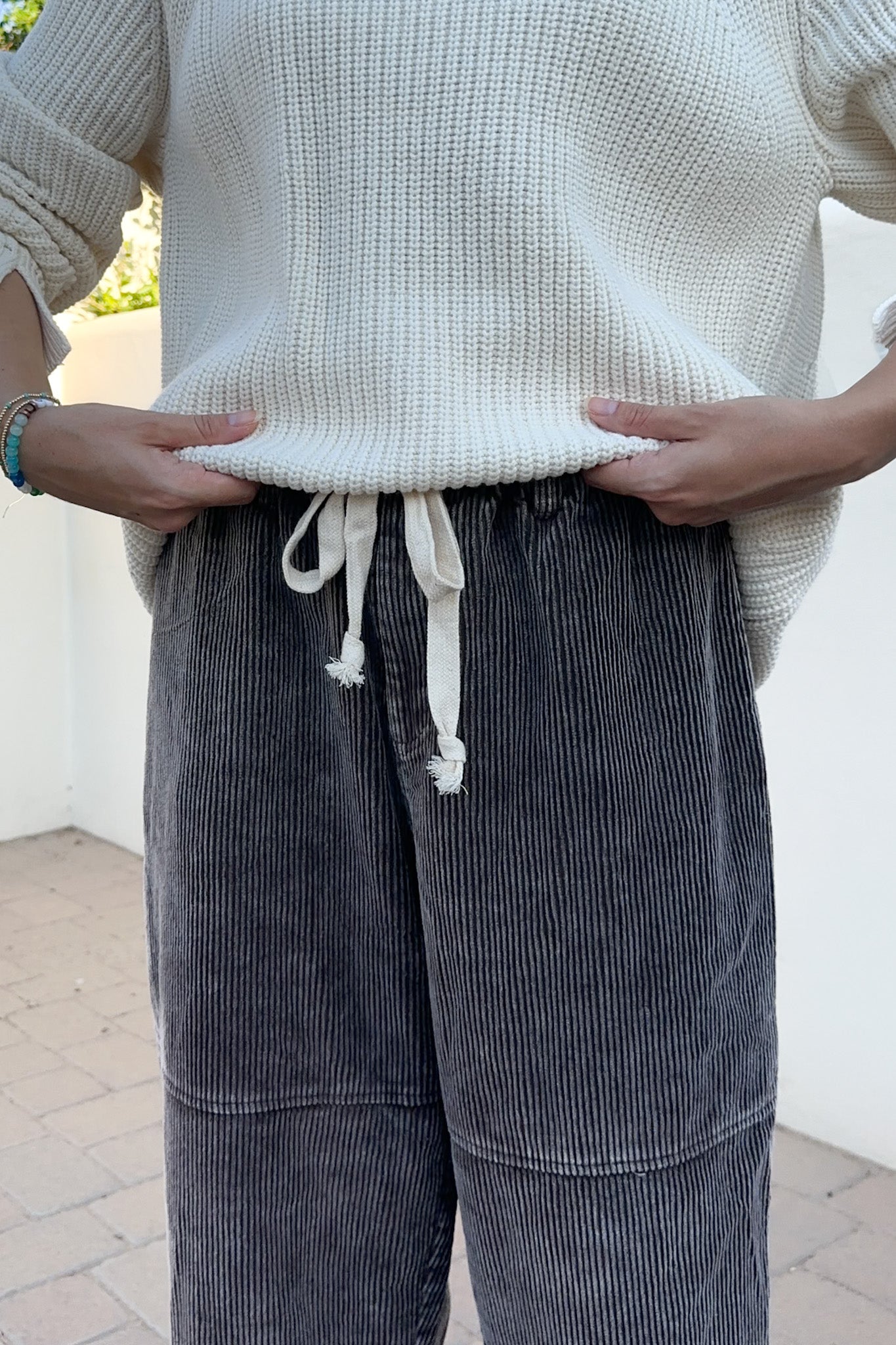 A person wearing dark gray corduroy wide-leg pants with an off-white drawstring and a cream ribbed knit sweater, standing outdoors against a white wall.